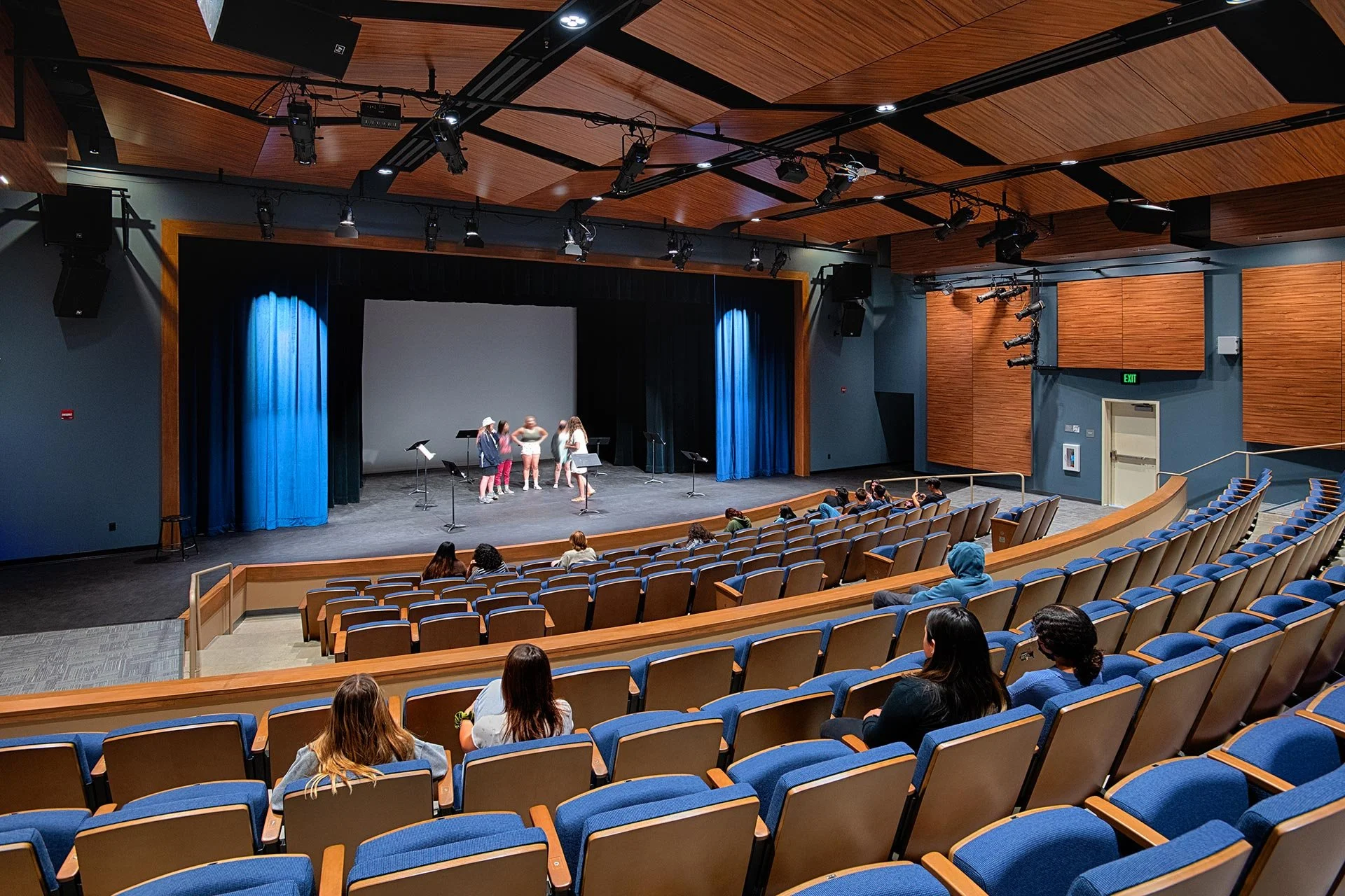 A theater with a stage where a group of children are rehearsing or performing. The auditorium has blue seats, wood-paneled walls, and ceiling, with overhead lighting and sound equipment. People are seated, watching the children.