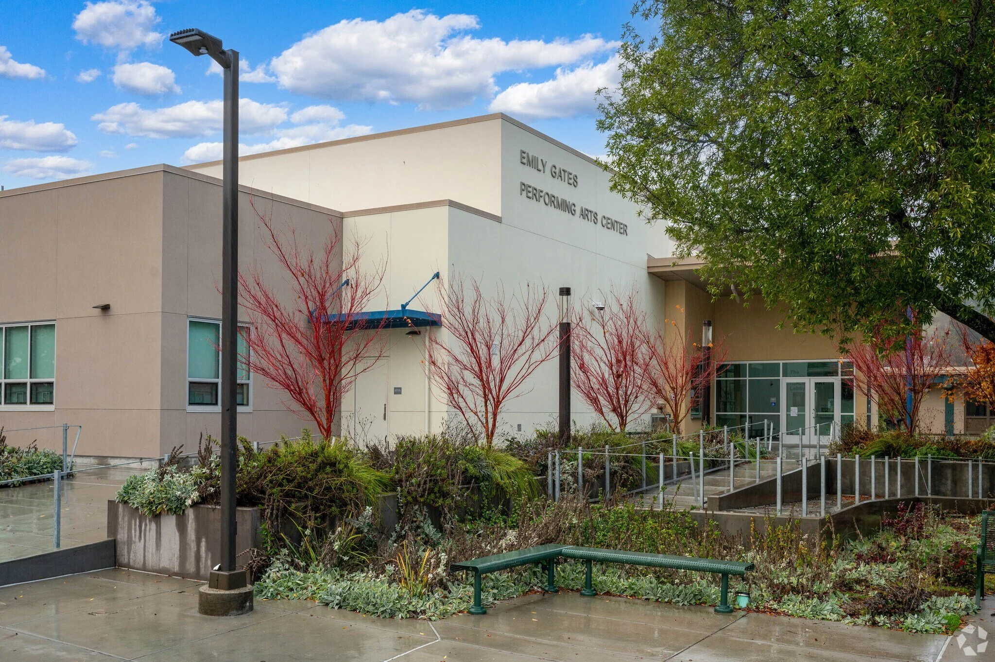 Exterior view of Emily Gates Performing Arts Center with trees, a lamppost, and a garden in the foreground on a cloudy day.