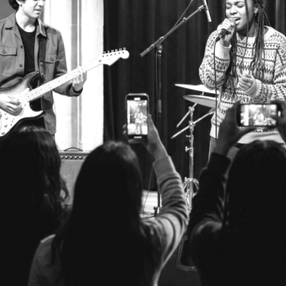 A woman singing into a microphone on stage with a guitarist next to her, while audience members record with their phones.