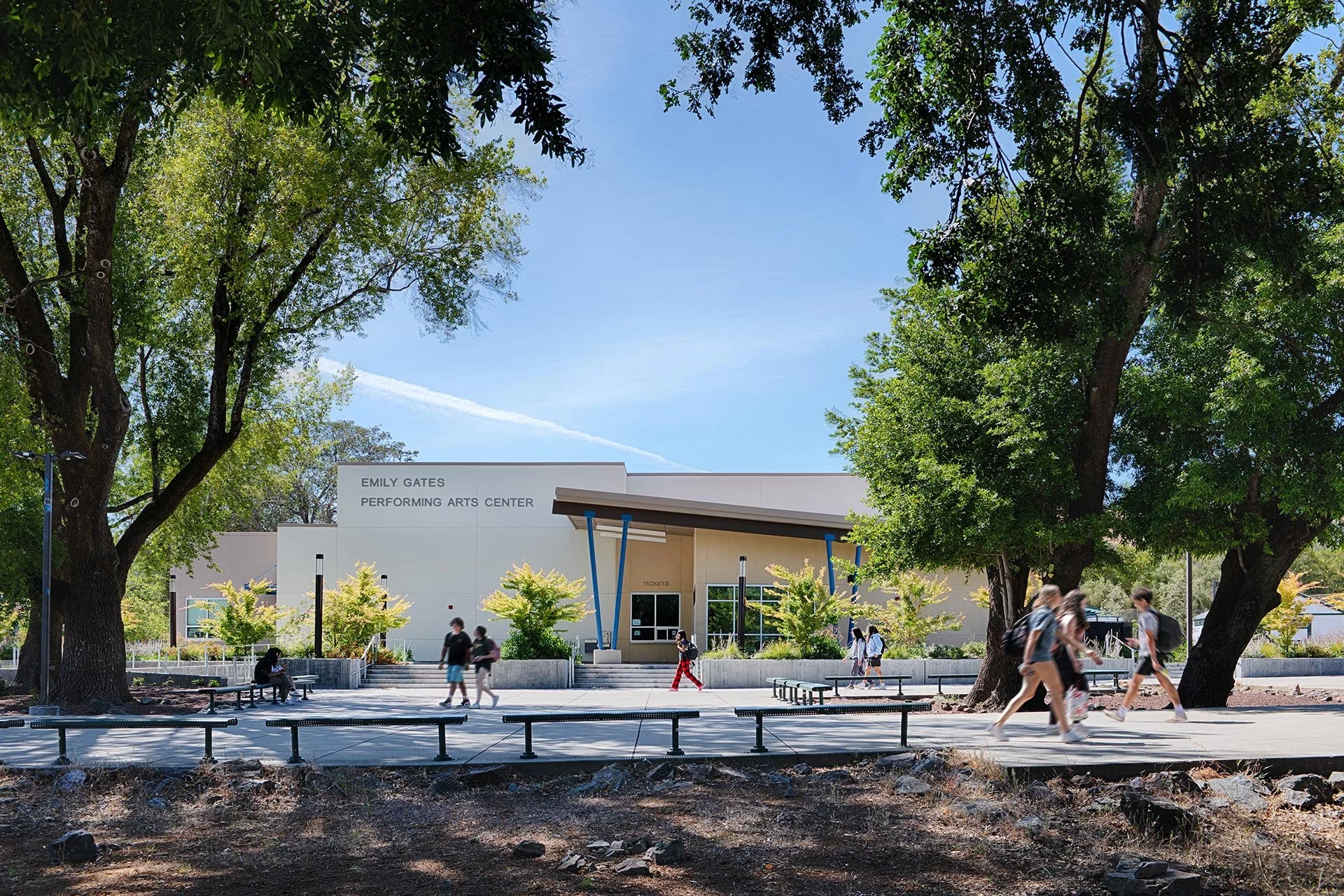 The Emily Gates Performing Arts Center with people walking and sitting outside, surrounded by large trees and a sunny sky.
