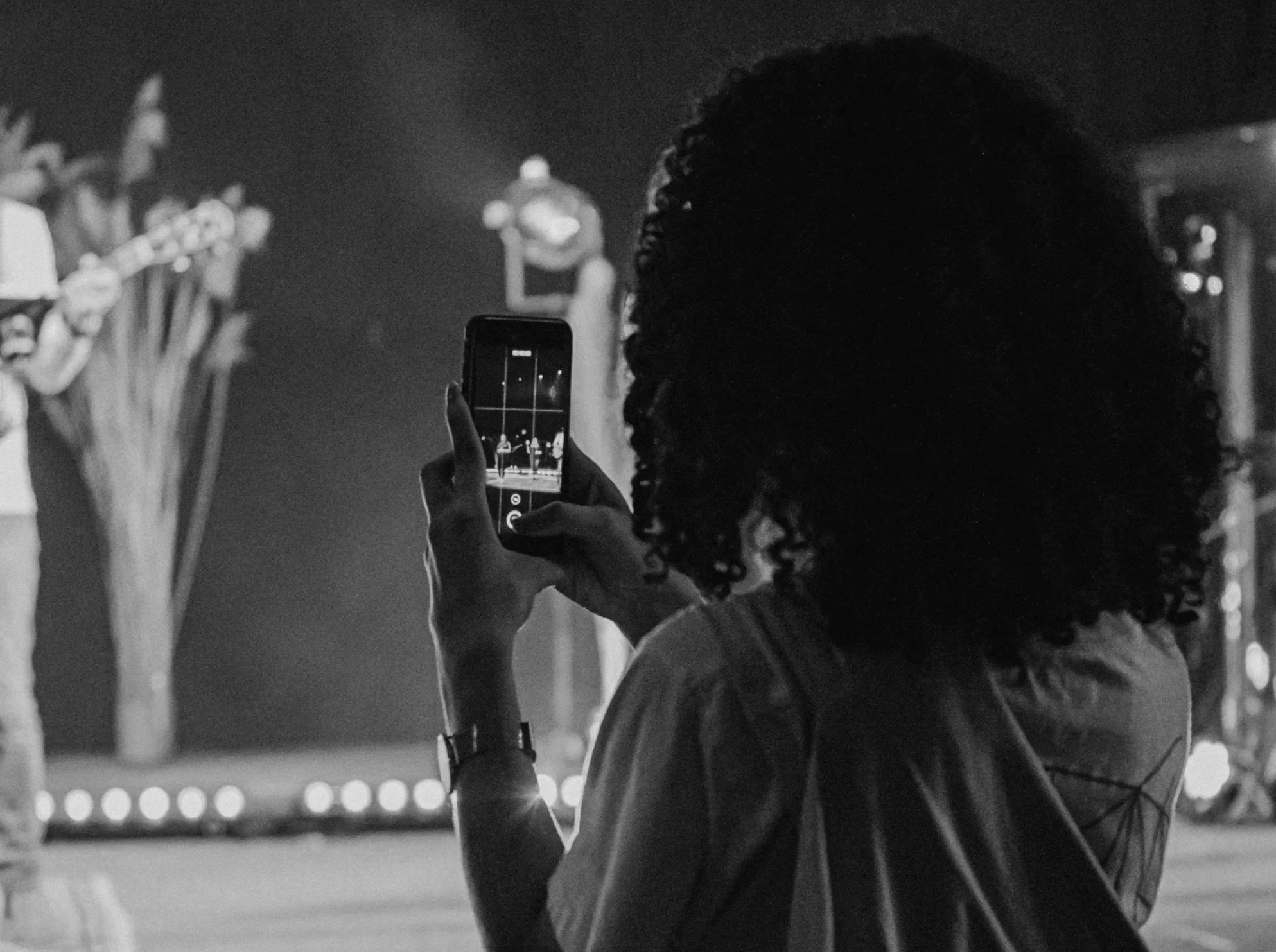 A person with curly hair is taking a photo or video of a stage performance with a smartphone. The performance features musicians and a singer, visible in the reflection of the person's phone screen, on stage with bright lights and blurred background.