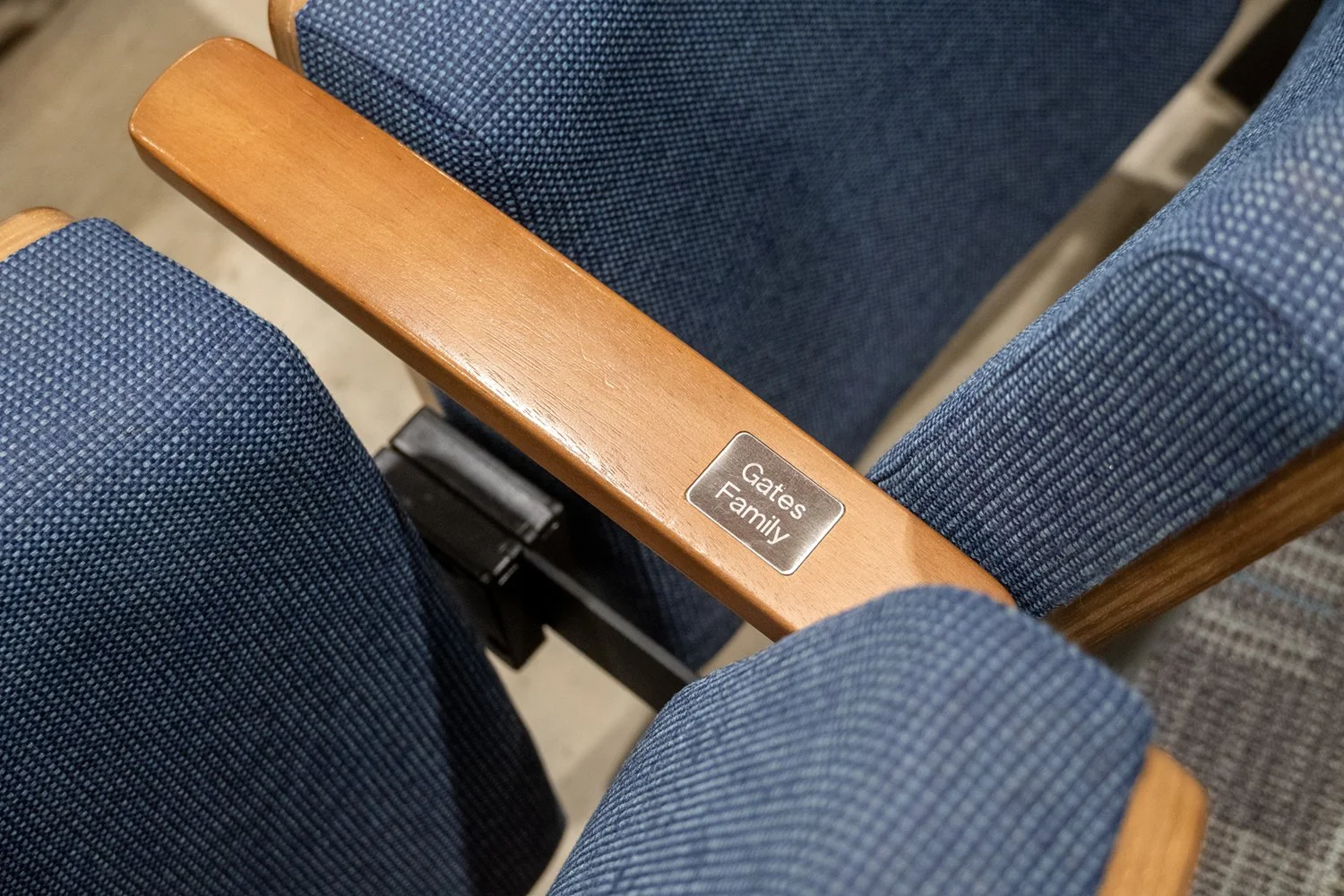 Blue theater or auditorium seats with wooden armrests, one armrest labeled 'Gates Family'.