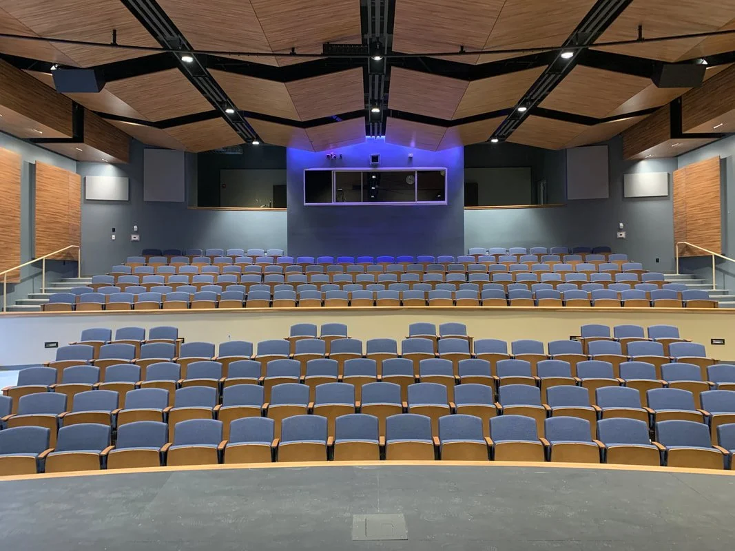 The Emily Gates Performing Arts Center  with rows of blue cushioned chairs, wooden accents on the walls and ceiling, and a large screen at the front with blue lighting.