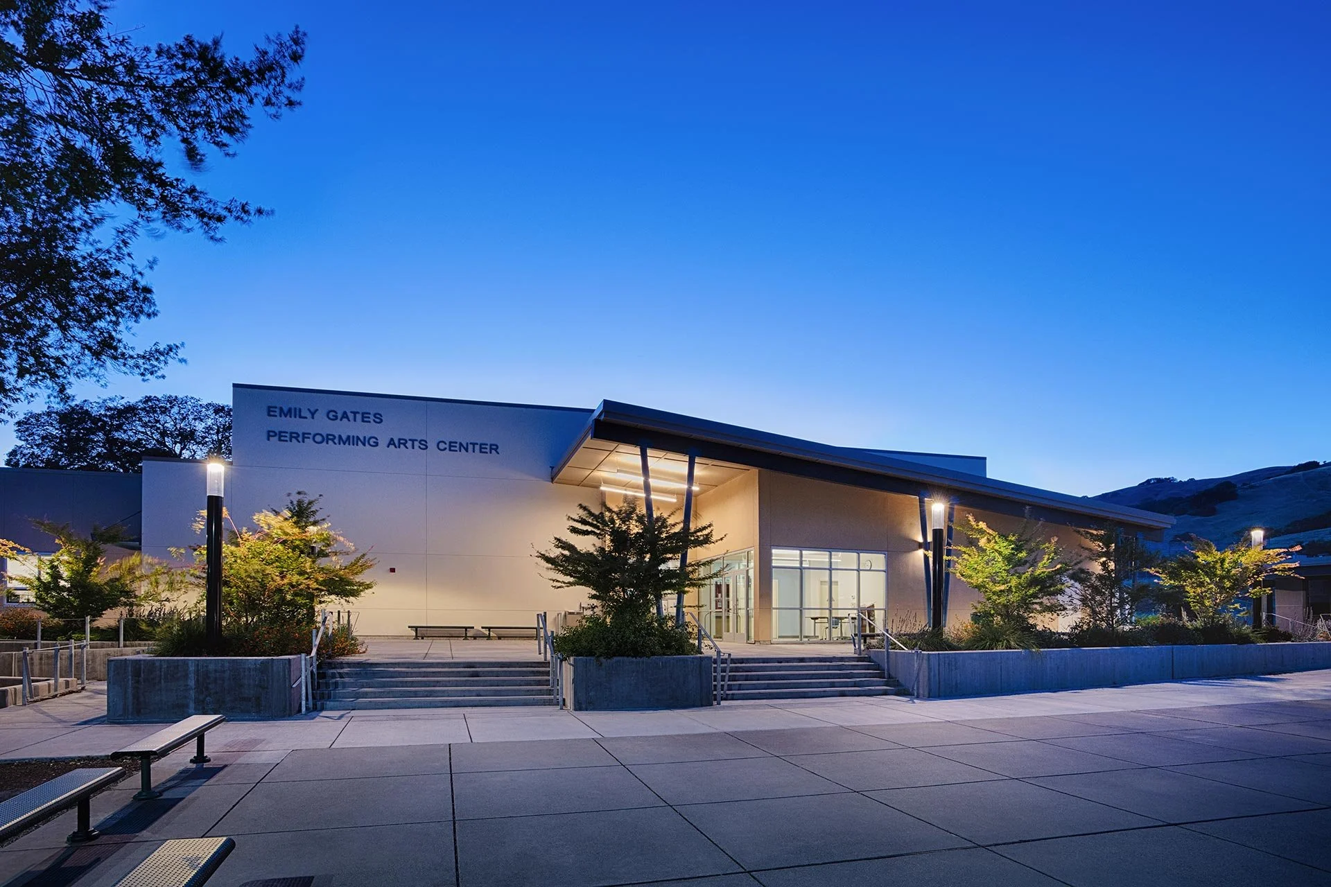 The Emily Gates Performing Arts Center at dusk, with exterior lighting, trees, benches, and steps leading to glass entrance doors.