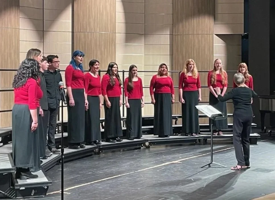 Group of young high school students singing on a stage, directed by a conductor, in a concert hall.