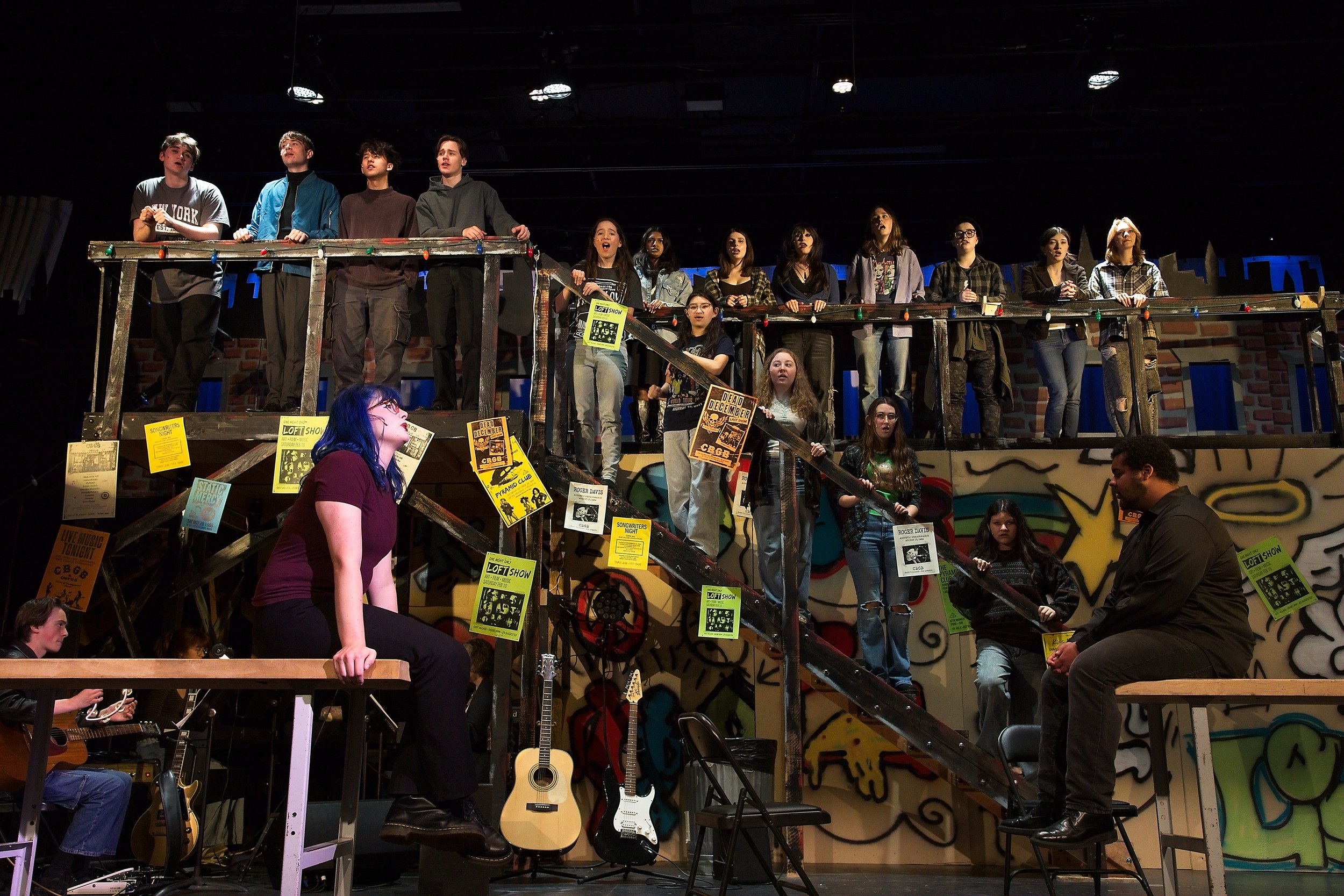 Stage performance with multiple people standing and sitting on a multi-level wooden structure. Musicians with guitars below, colorful posters on the structure, a dark background, and a graffiti-style mural on the wall.