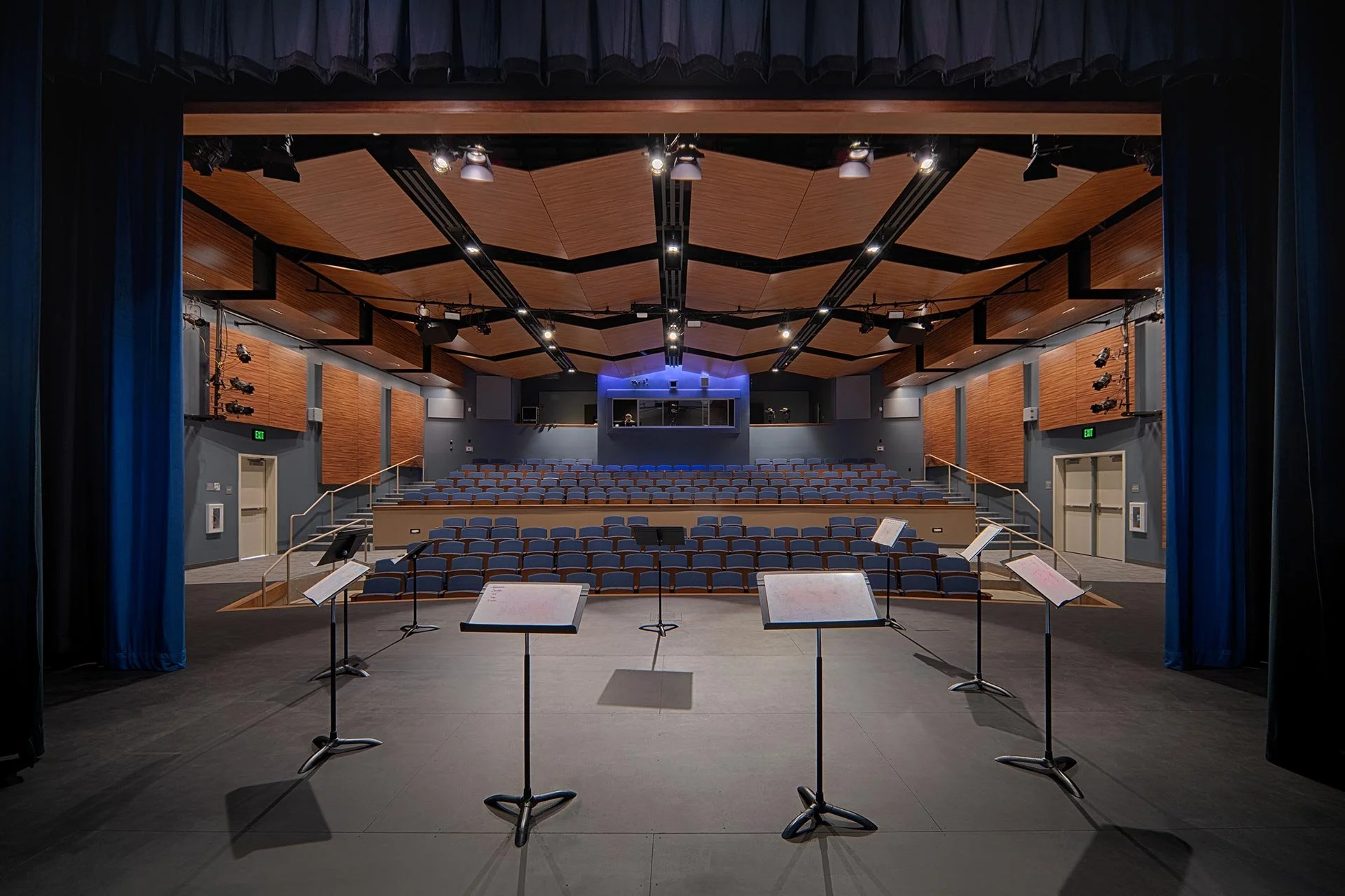 The Emily Gates Performing Arts Center stage with music stands and chairs, view from the stage looking out towards rows of seats and a balcony with sound and lighting equipment, blue curtains framing the stage.
