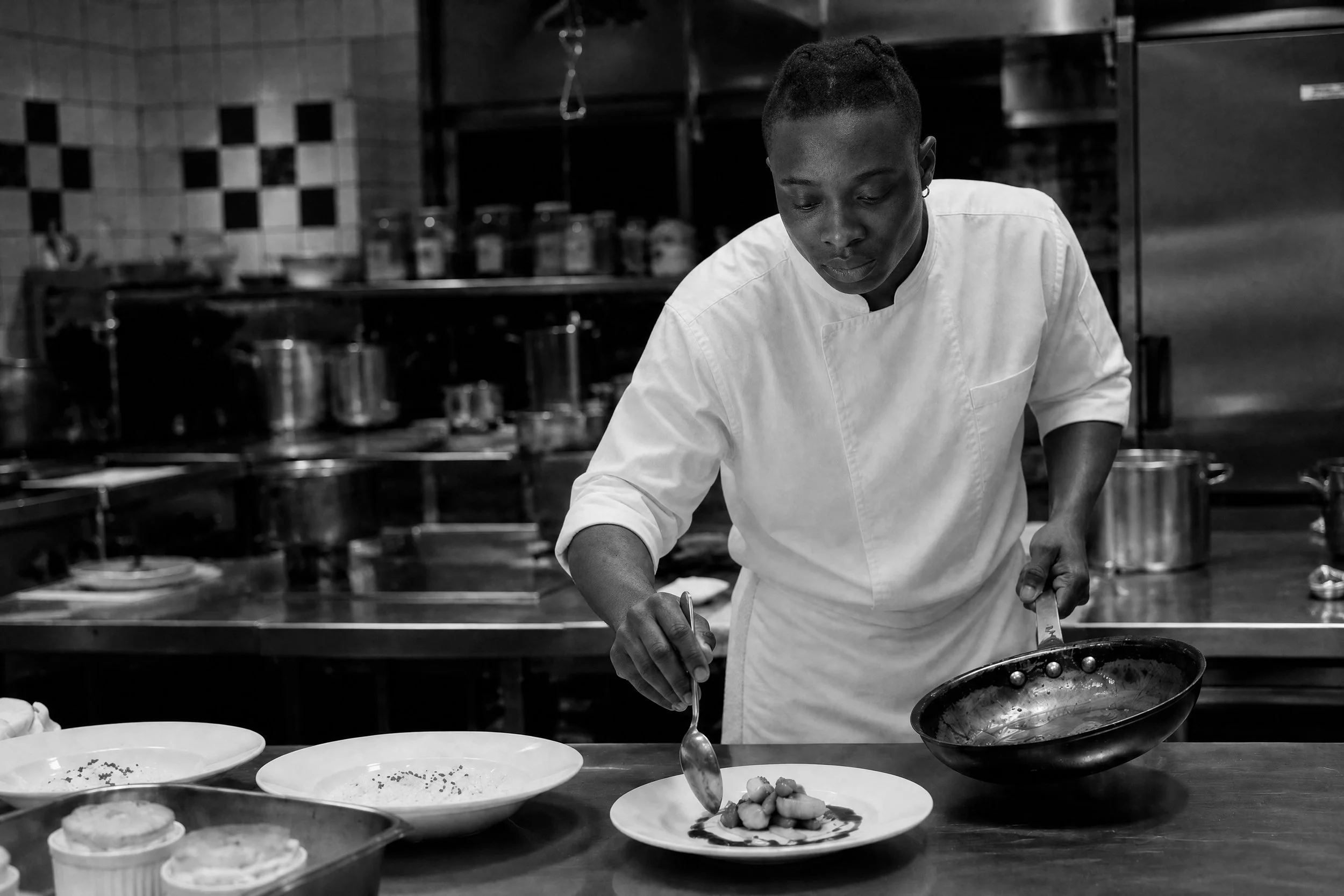 A chef in ChefShield™ chef jacket plates food in a professional kitchen with pots and utensils in the background.