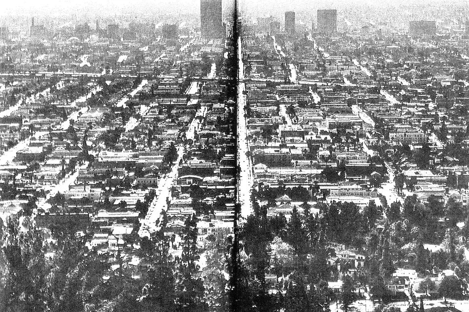A black and white aerial photograph of a city street grid, taken from a high vantage point, showing densely packed buildings and trees.