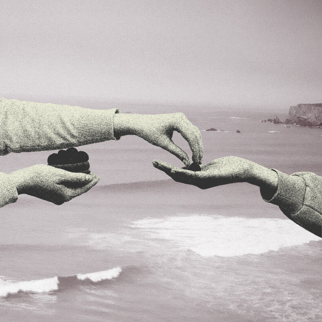 Black and white photo of two hands reaching towards each other over a beach with cliffs in the background.