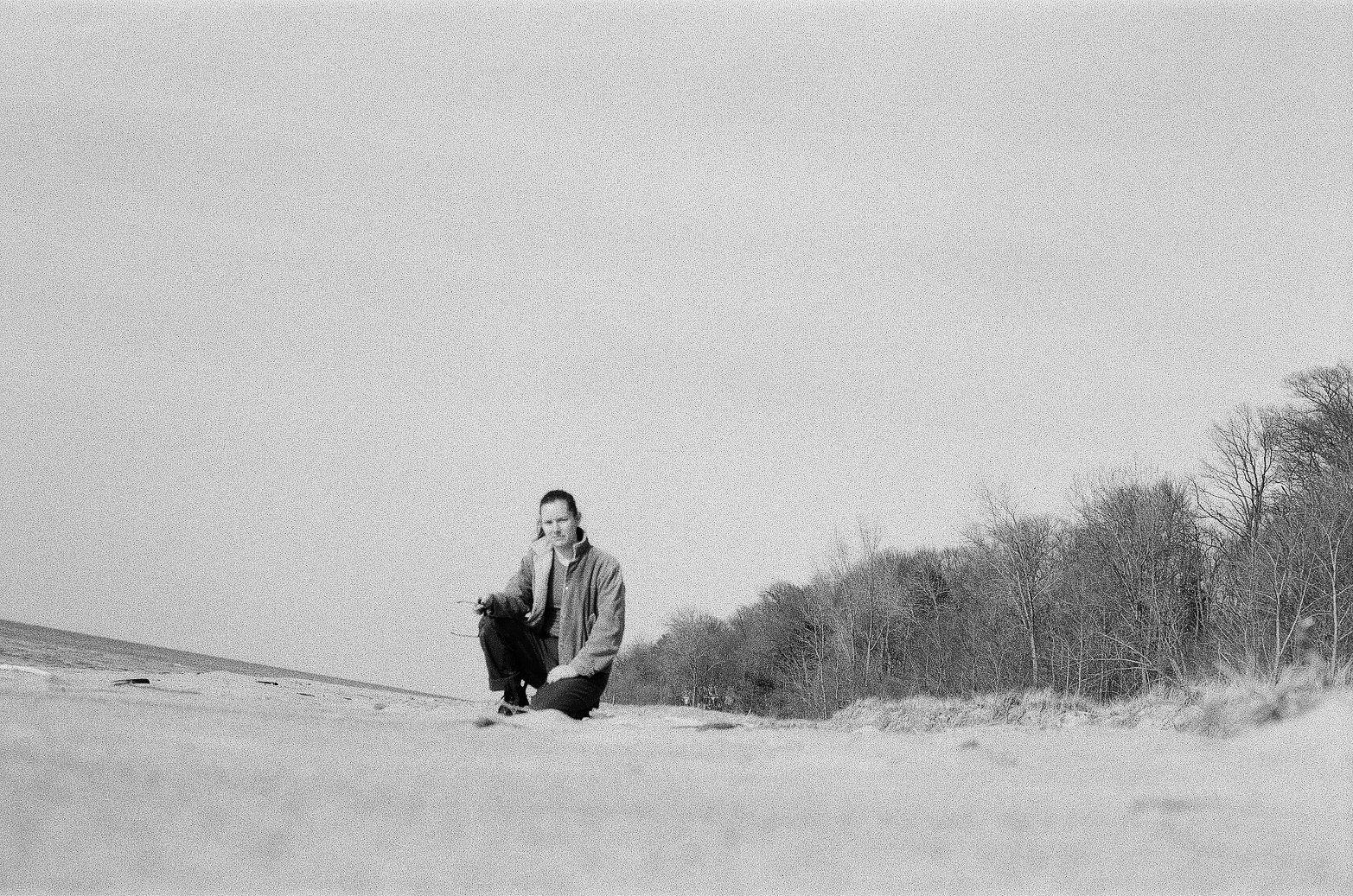 A person kneeling on the sand at the beach near trees with bare branches, under a clear sky.