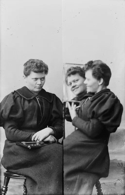 Black and white photo of three young girls seated on stools, with two of them looking at a small mirror and smiling, while the girl on the left looks serious.