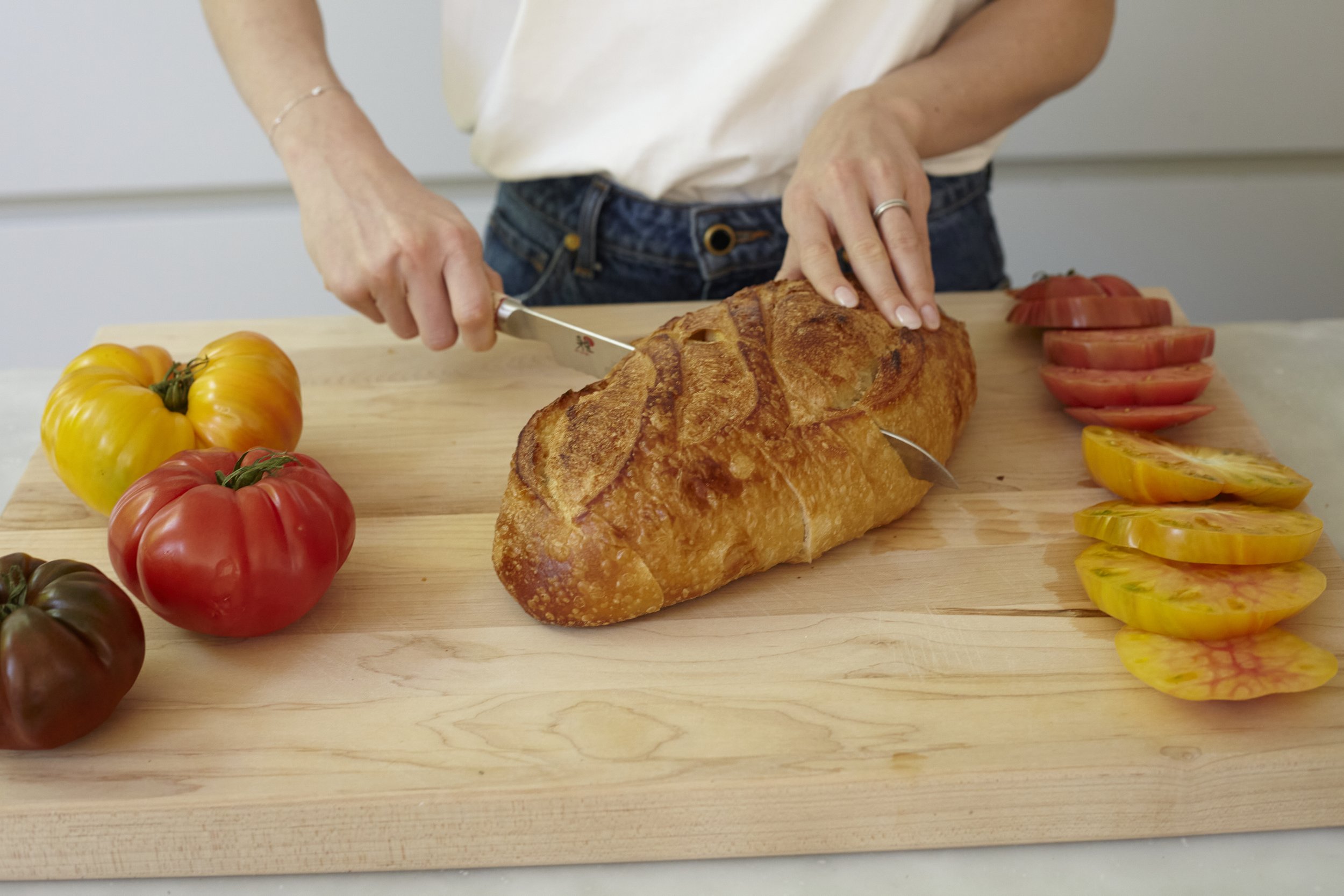 A person slicing a large loaf of bread on a wooden cutting board surrounded by colorful heirloom tomatoes, some sliced, in a kitchen setting.