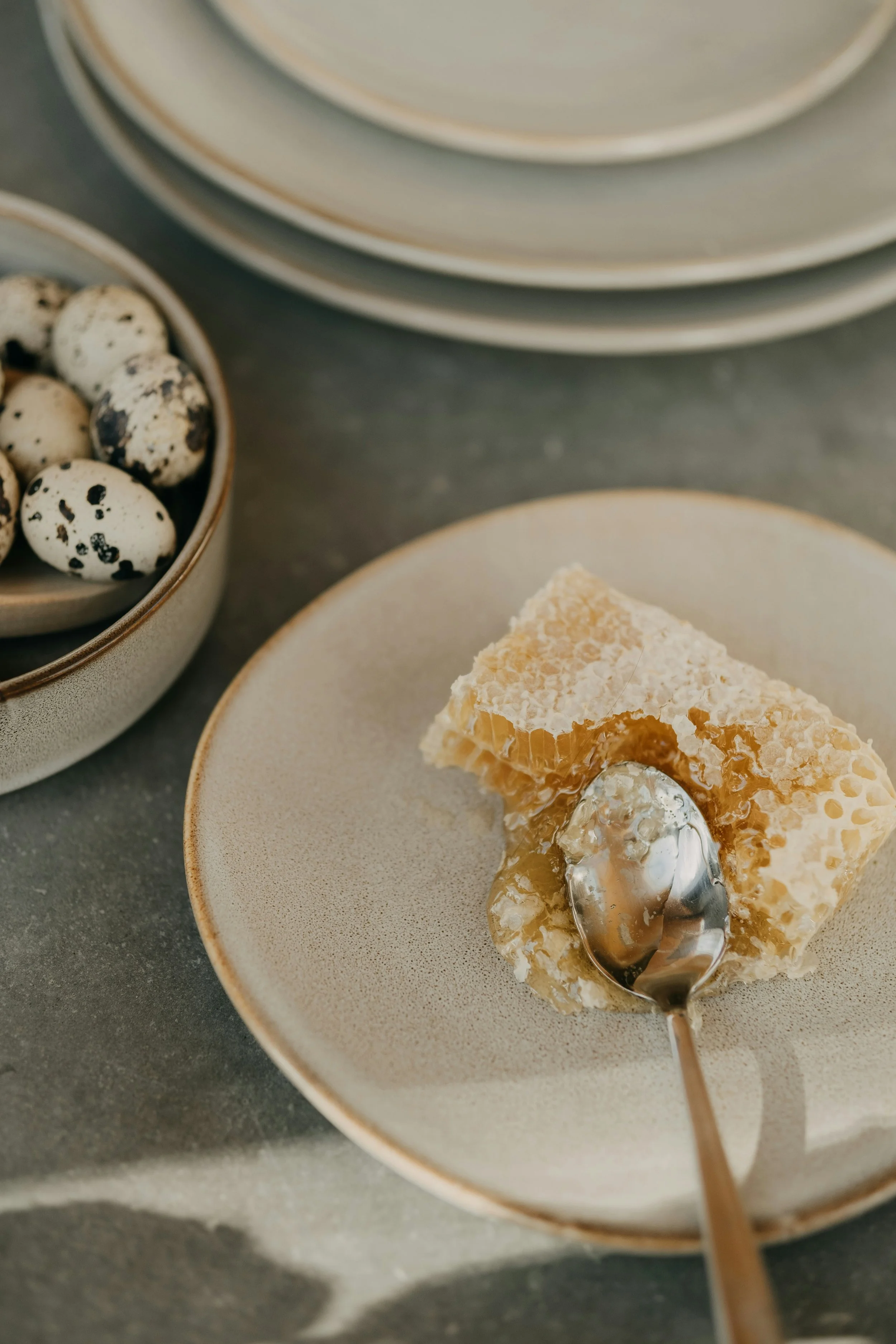 Close-up of a honeycomb piece on a plate with a silver spoon, with a bowl of quail eggs in the background.