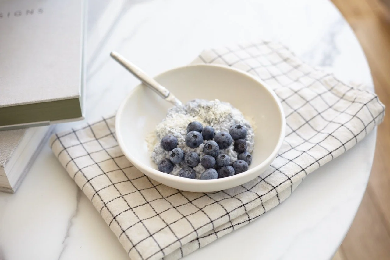 A bowl of blueberries with powdered sugar and a spoon on a checkered cloth next to a stack of books.