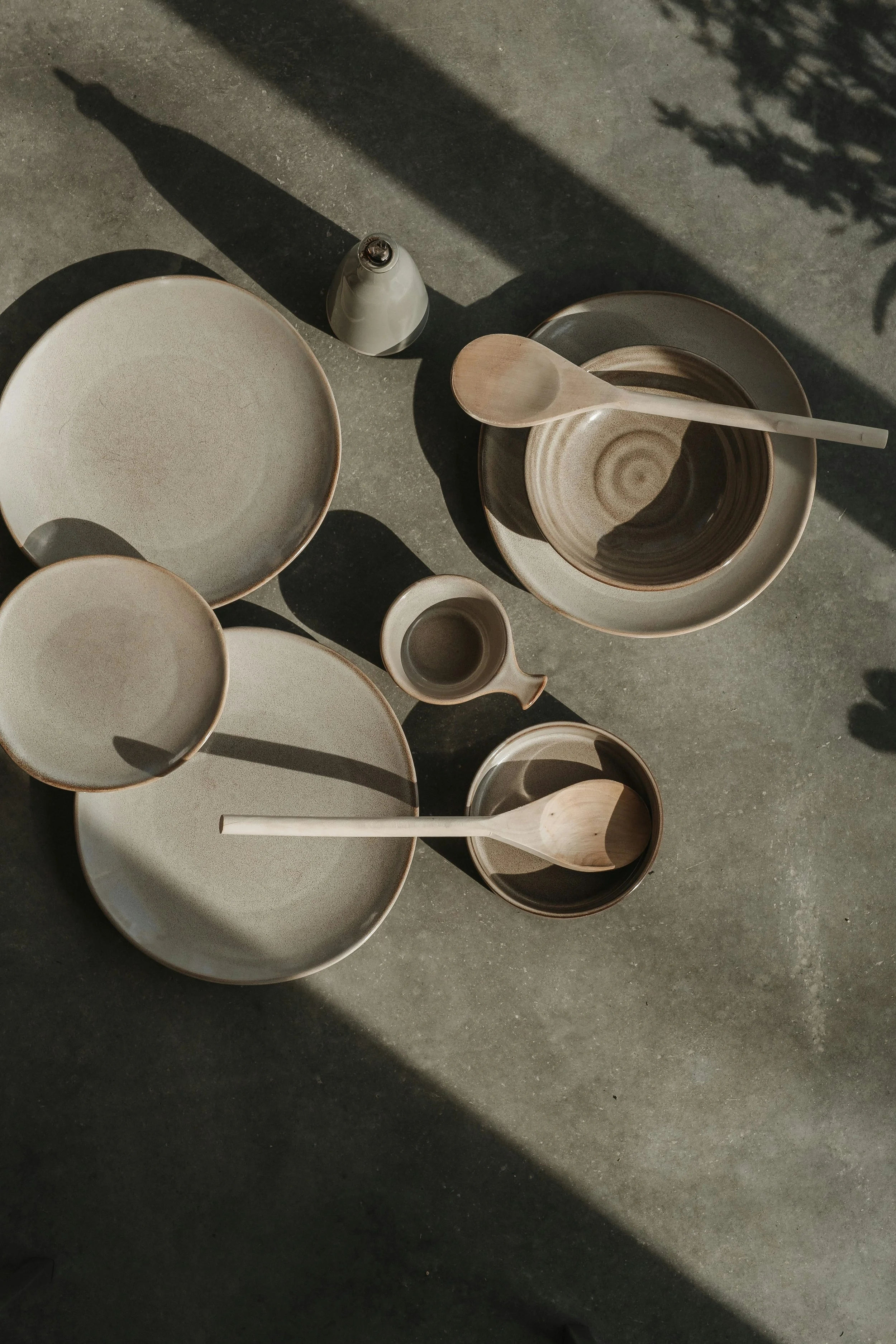 Empty ceramic bowls, a wooden spoon, a small pitcher, and plates on a textured gray surface with shadows.