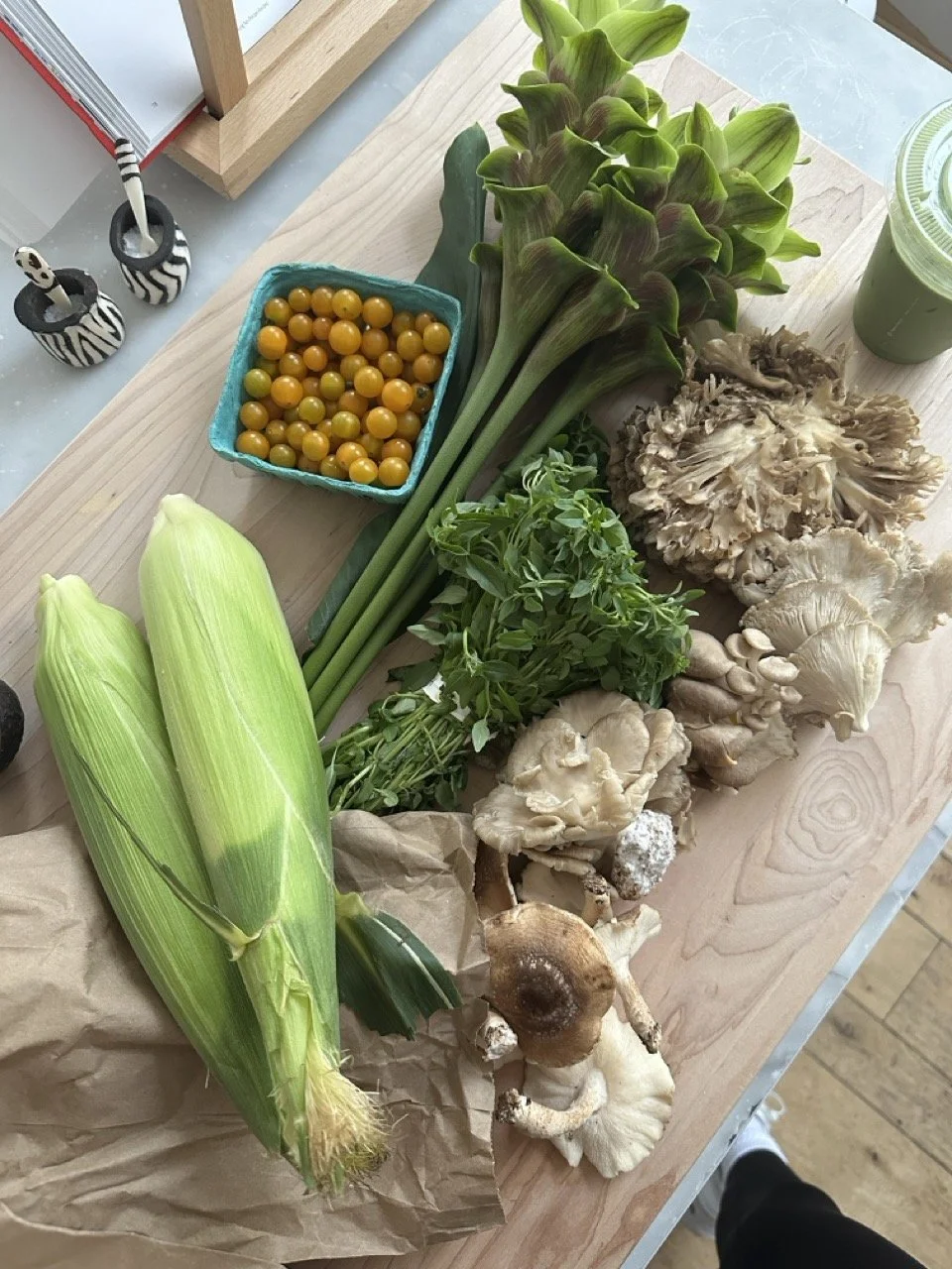 A variety of fresh vegetables and mushrooms on a wooden table, including corn, okra, greens, and different types of mushrooms.
