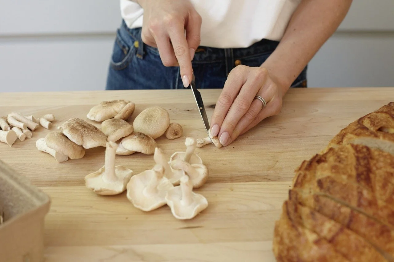Person slicing a mushroom on a wooden table, surrounded by various mushrooms and a pie.