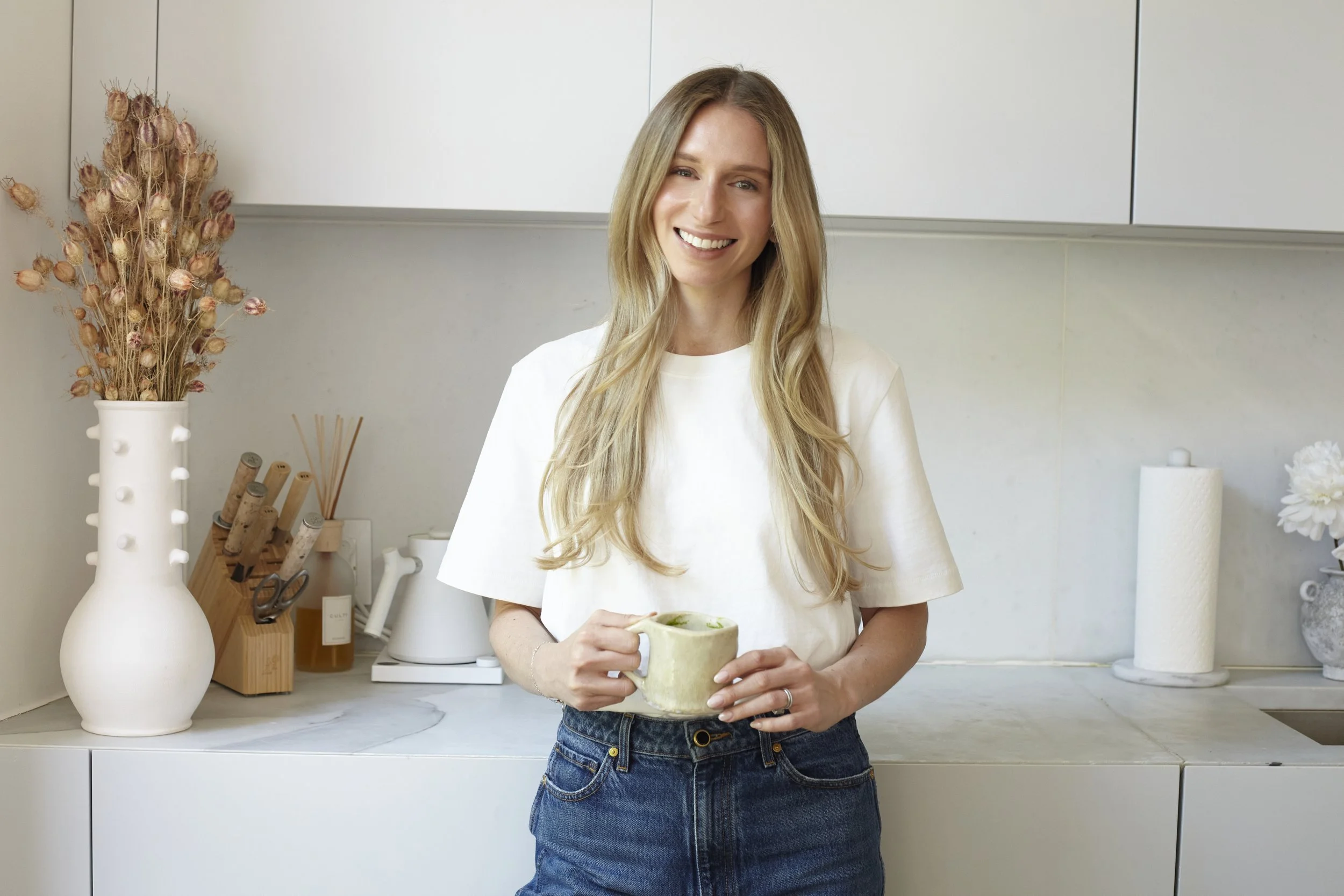 A woman with long blonde hair smiling and holding a beige ceramic mug in a modern kitchen.