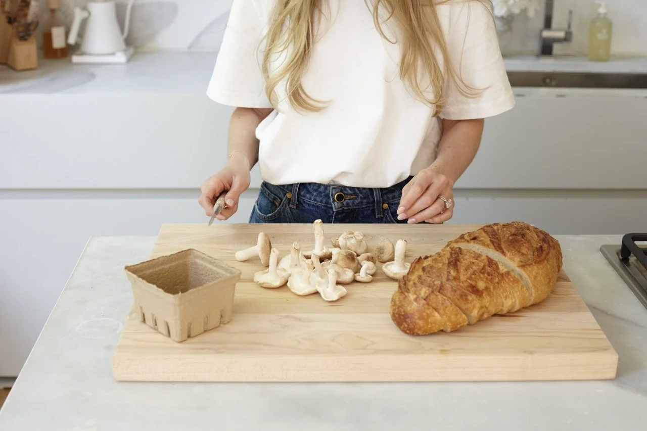 Person slicing mushrooms on a wooden cutting board with a loaf of bread nearby in a modern kitchen.