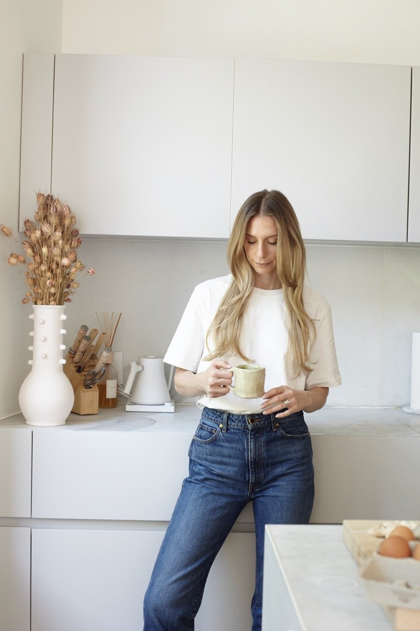 A woman with long blonde hair wearing a white t-shirt and blue jeans holding a mug in a modern, white kitchen with minimal decor