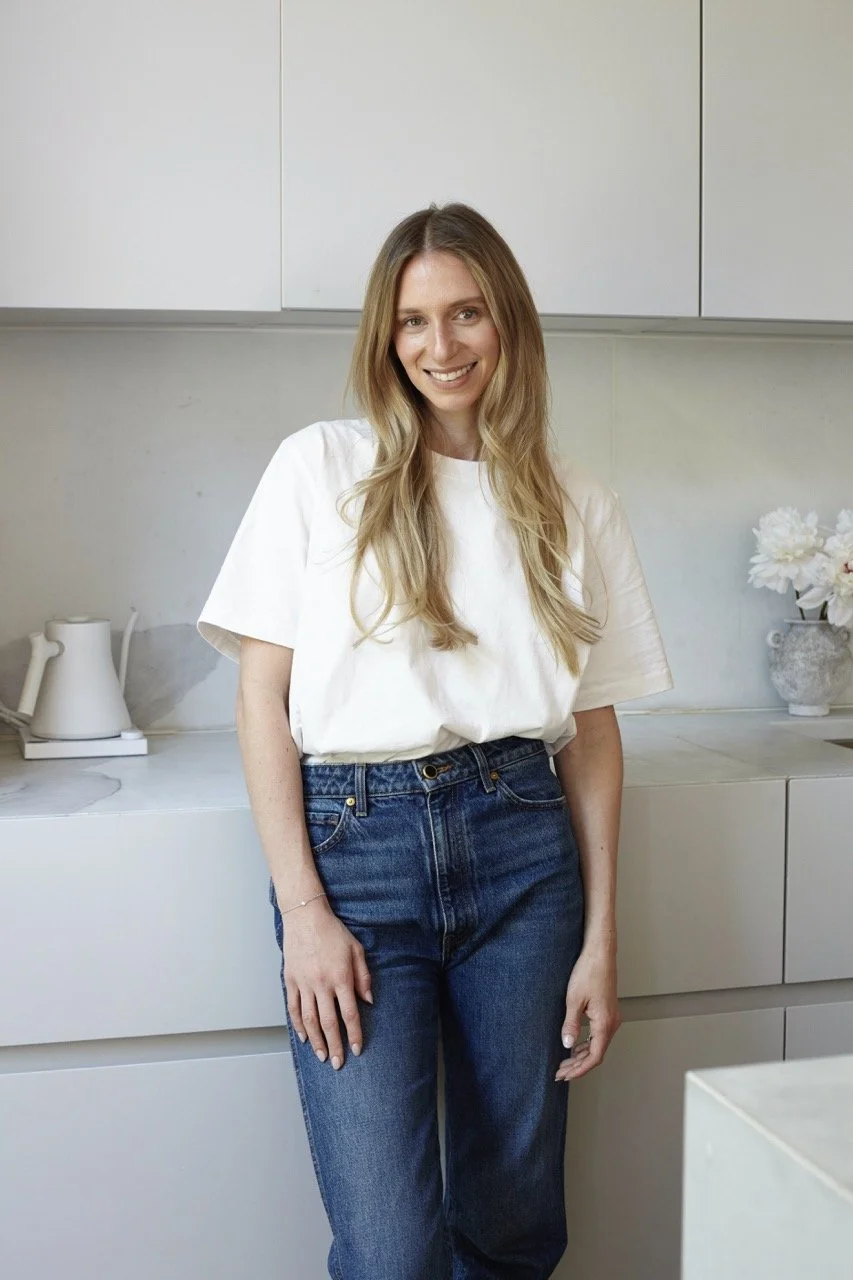 A woman with long blonde hair, smiling, wearing a white t-shirt and blue jeans, standing in a modern kitchen with white cabinets and flowers in a pot on the counter.