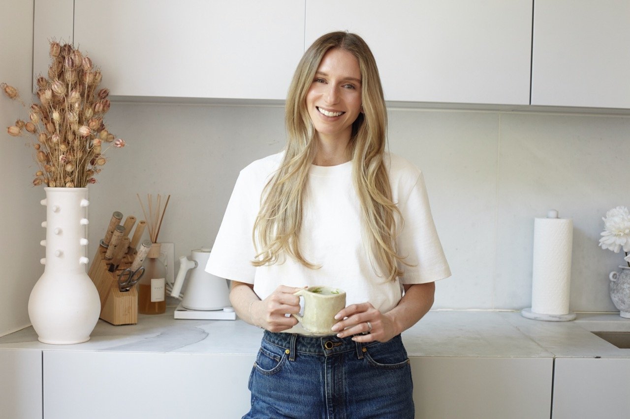 A woman with long blonde hair smiling and holding a mug in a modern kitchen with white cabinets, dried flowers in a white vase, and kitchen utensils on the counter.
