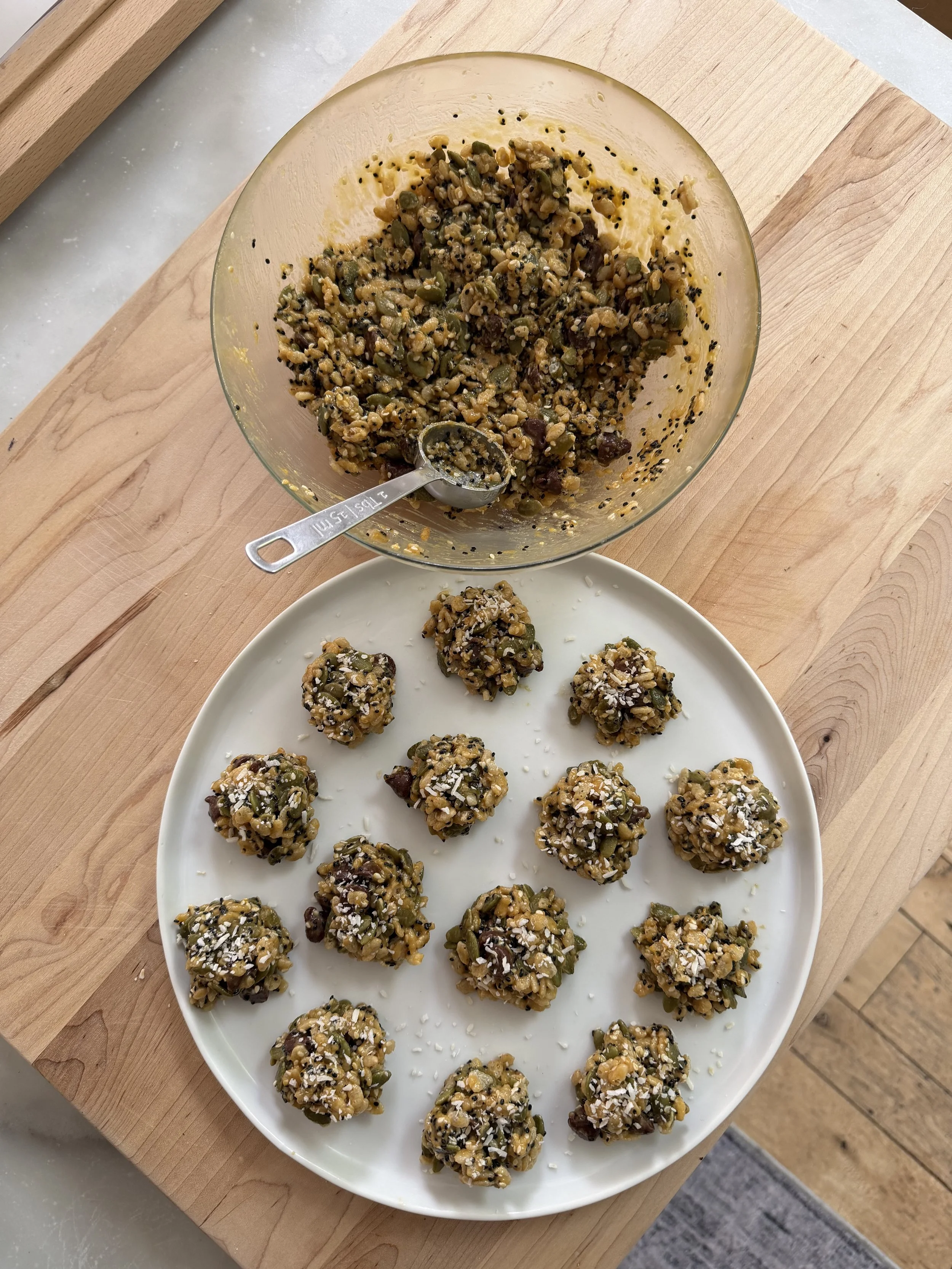 A white plate with homemade energy bites made from oats, seeds, nuts, and dried fruit, sprinkled with shredded coconut. The plate is on a wooden cutting board, with more bites and a mixing bowl in the background.