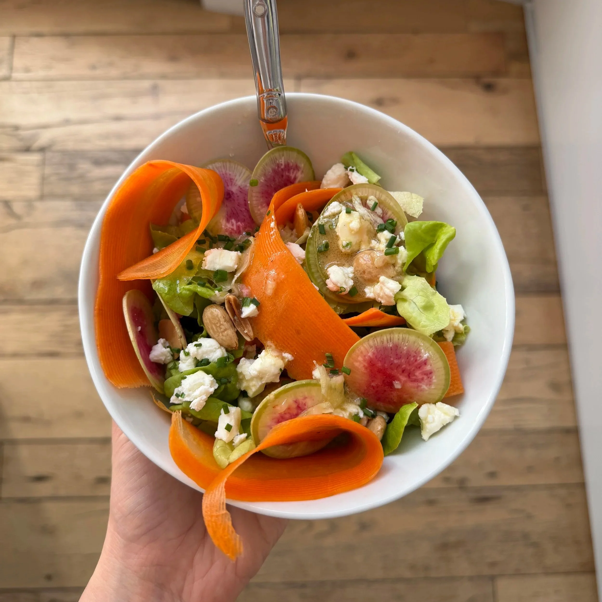 A hand holding a white bowl of sliced carrots, radishes, lettuce, and crumbled cheese, with a silver spoon inside, set against a wooden floor background.