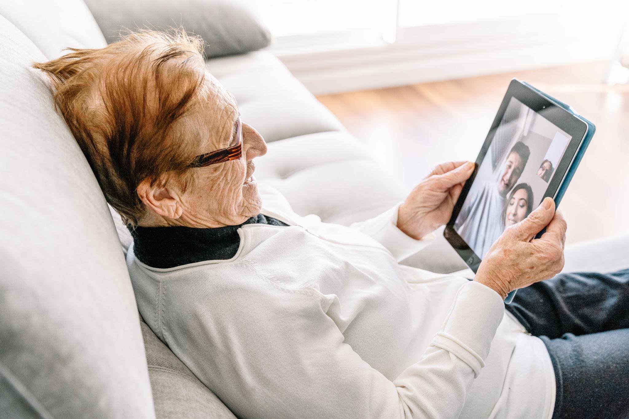 Picture of older woman relaxed on her couch talking to her grandchildren via a video on her tablet.