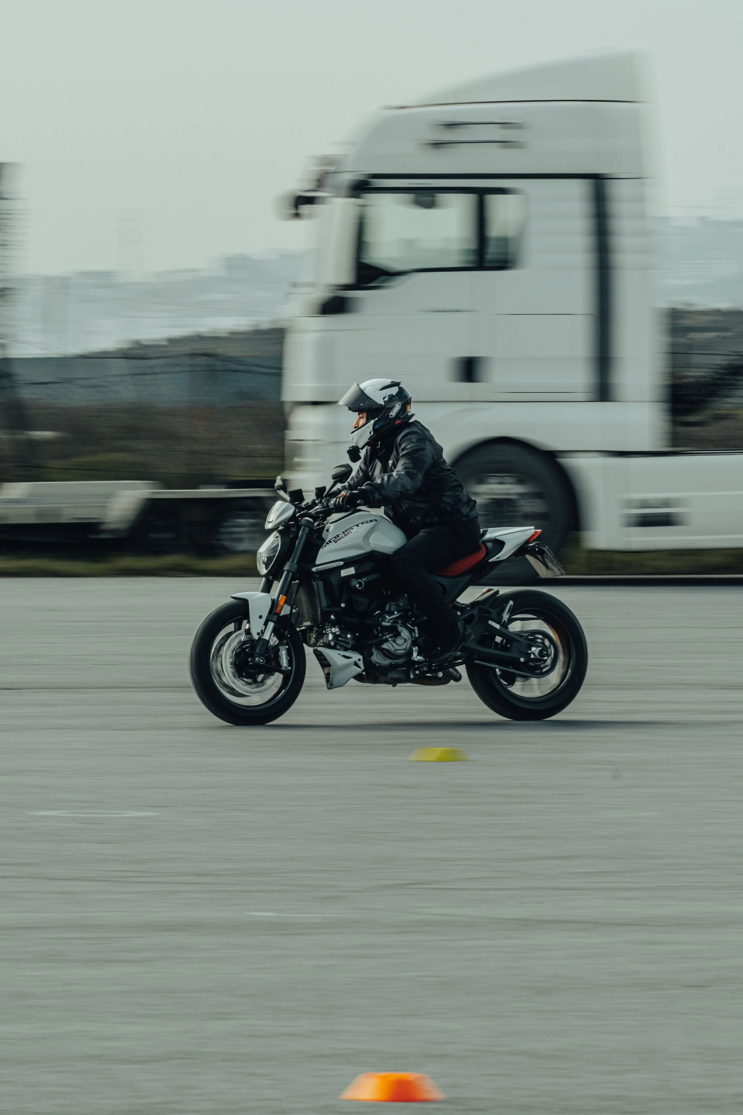 Motorcycle rider in helmet and jacket riding on open road, blurred background with large white truck and orange cone in foreground, overcast sky.
