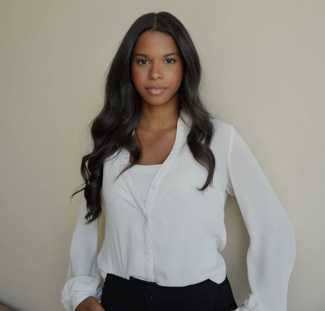 A young woman with long wavy black hair, wearing a white blouse and black pants, standing against a plain beige wall.
