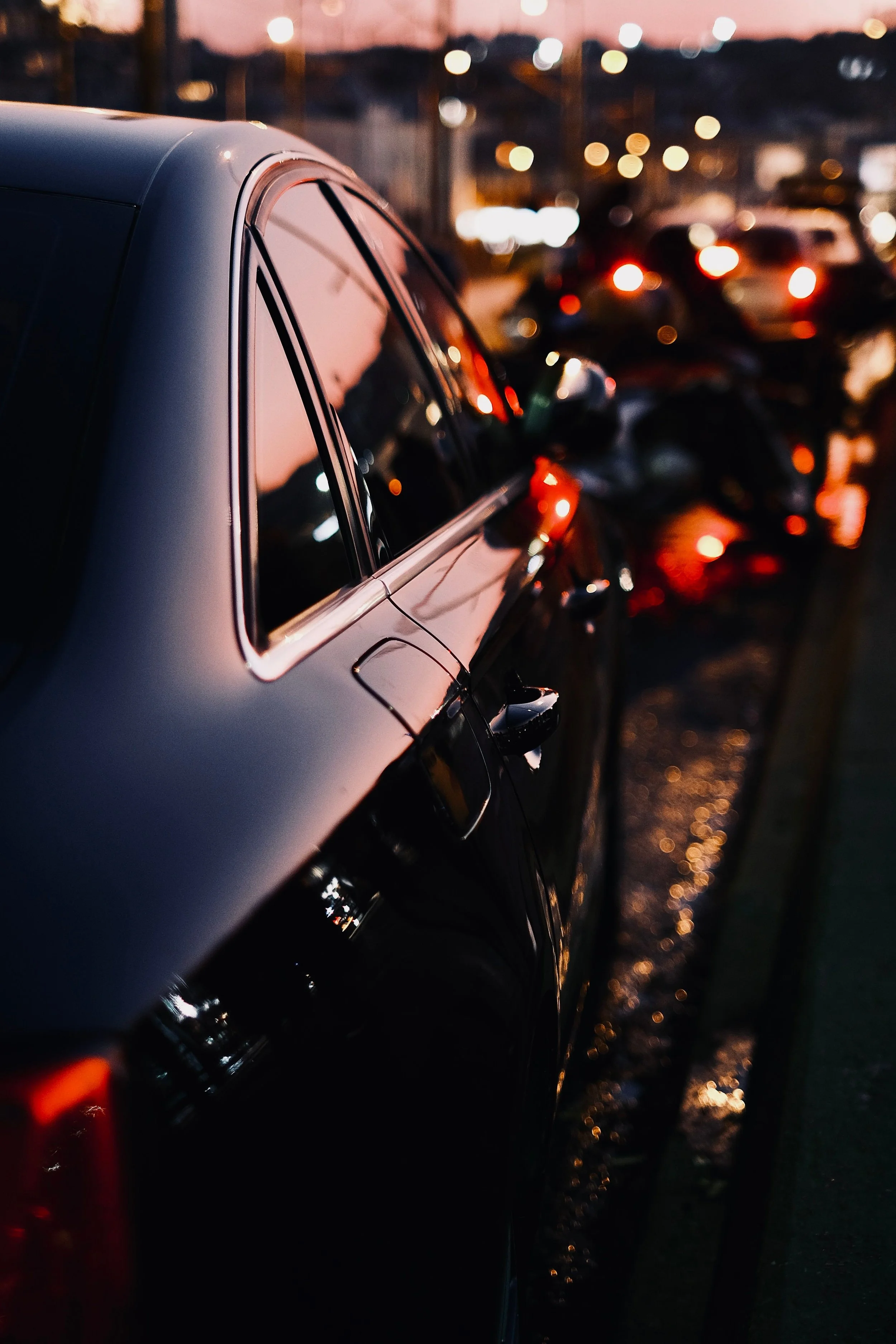 Line of parked cars on a wet street during twilight, with blurry city lights in the background and reflections on the ground.