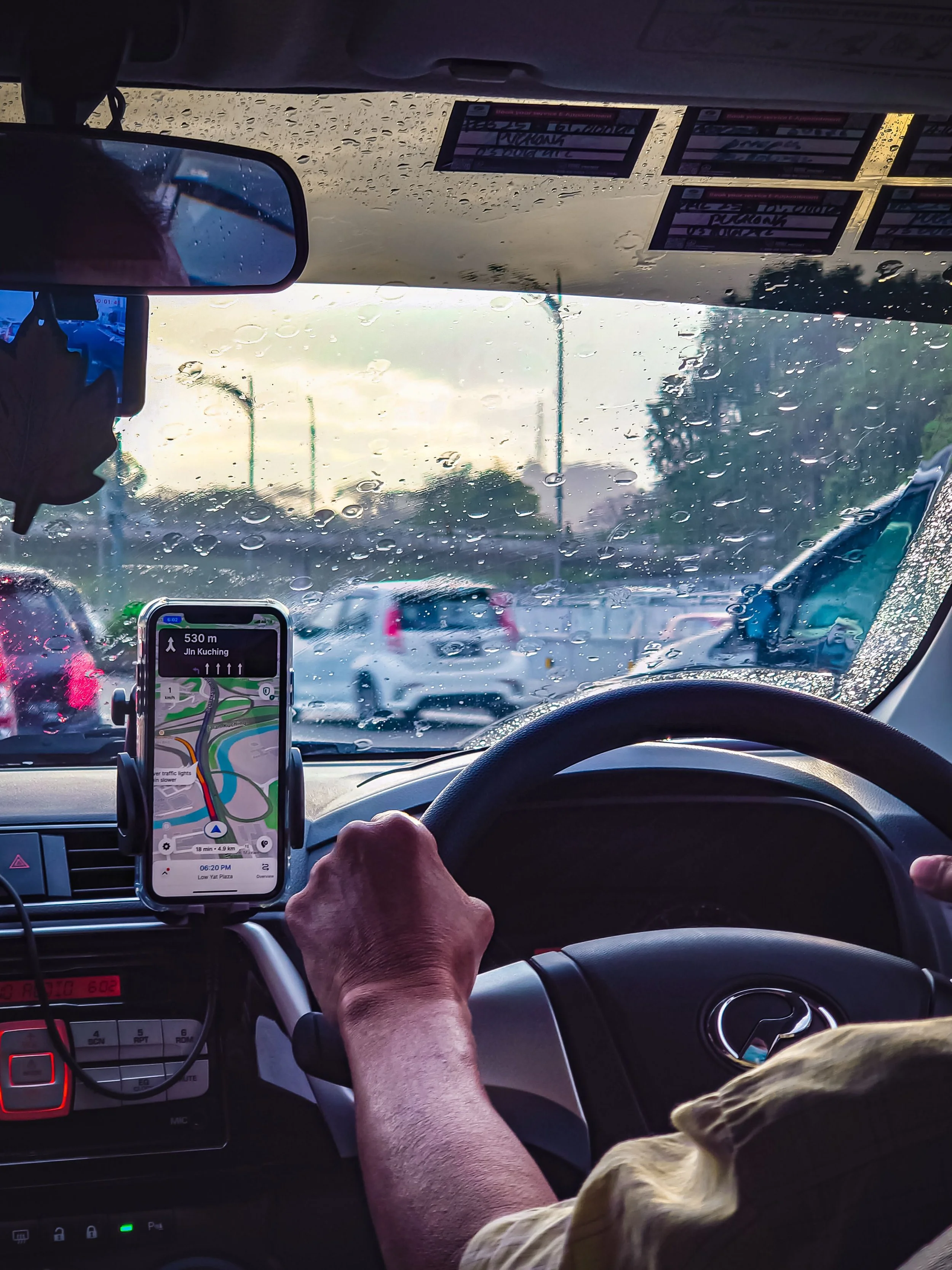 Inside a vehicle during rainy weather, showing a rearview mirror, a GPS device on the dashboard displaying directions to Jln Kuching, and an arm and dog resting on the seat. Raindrops are visible on the windshield.