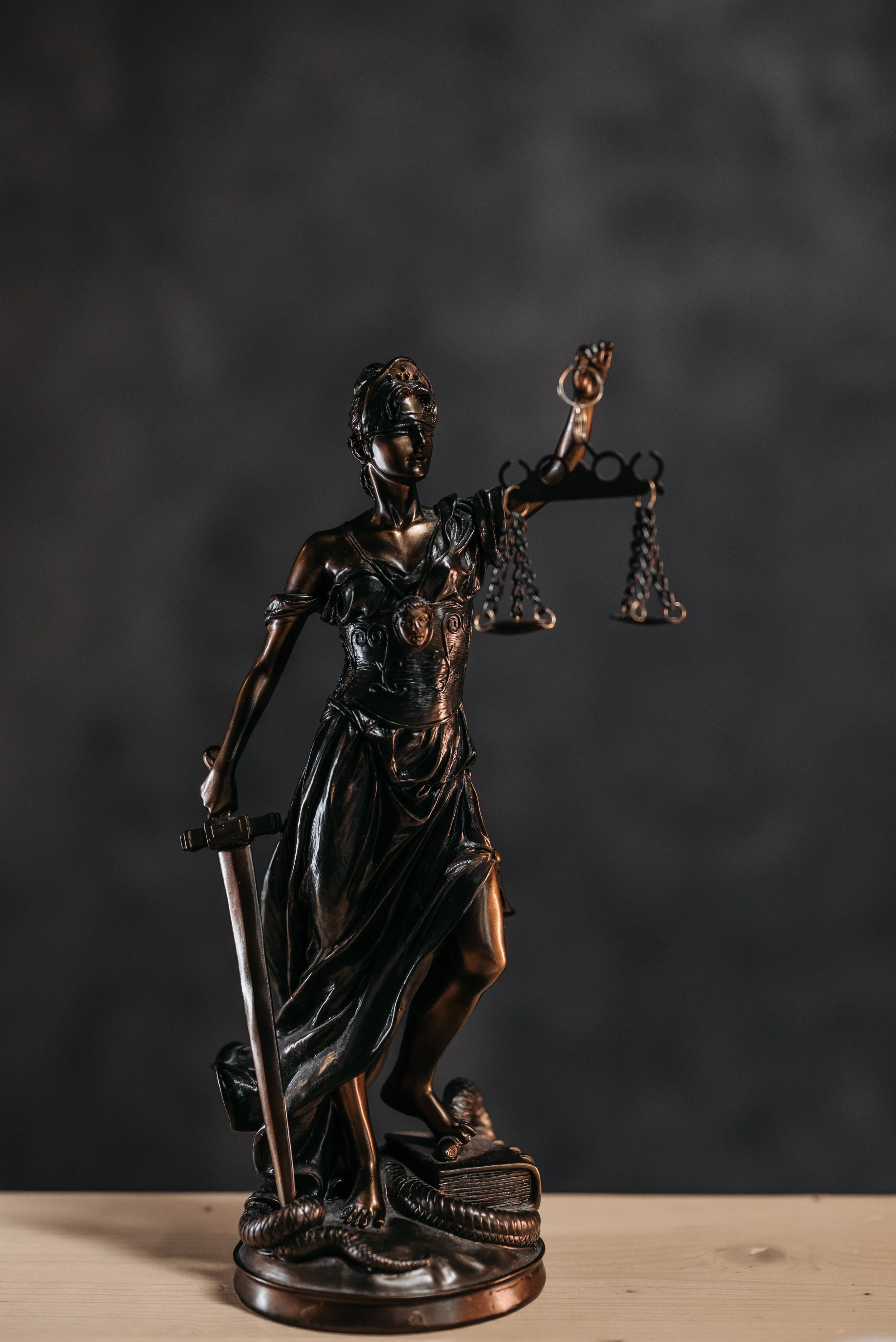 A bronze statue of Lady Justice holding a scale in her left hand and a sword in her right hand, standing on a base with a snake coiled around her foot.