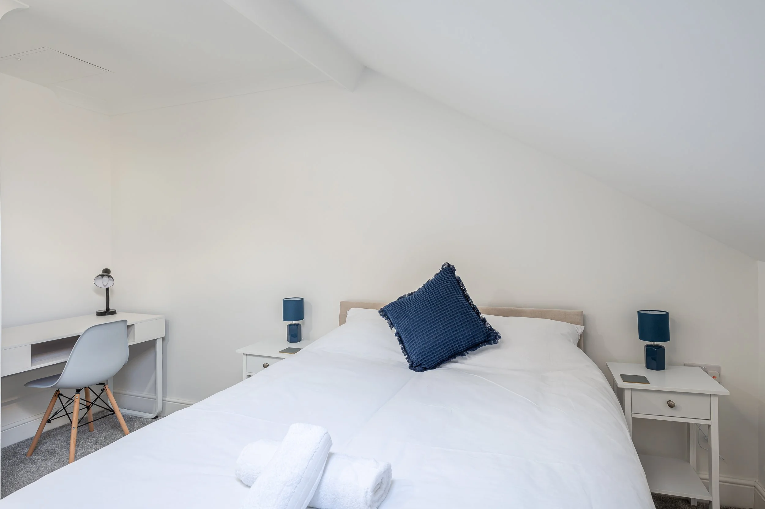 A bedroom with a white bed featuring a dark blue textured pillow, bedside tables with dark blue lamps, a white desk with a white chair, and a small lamp. The room has plain white walls and an angled ceiling.