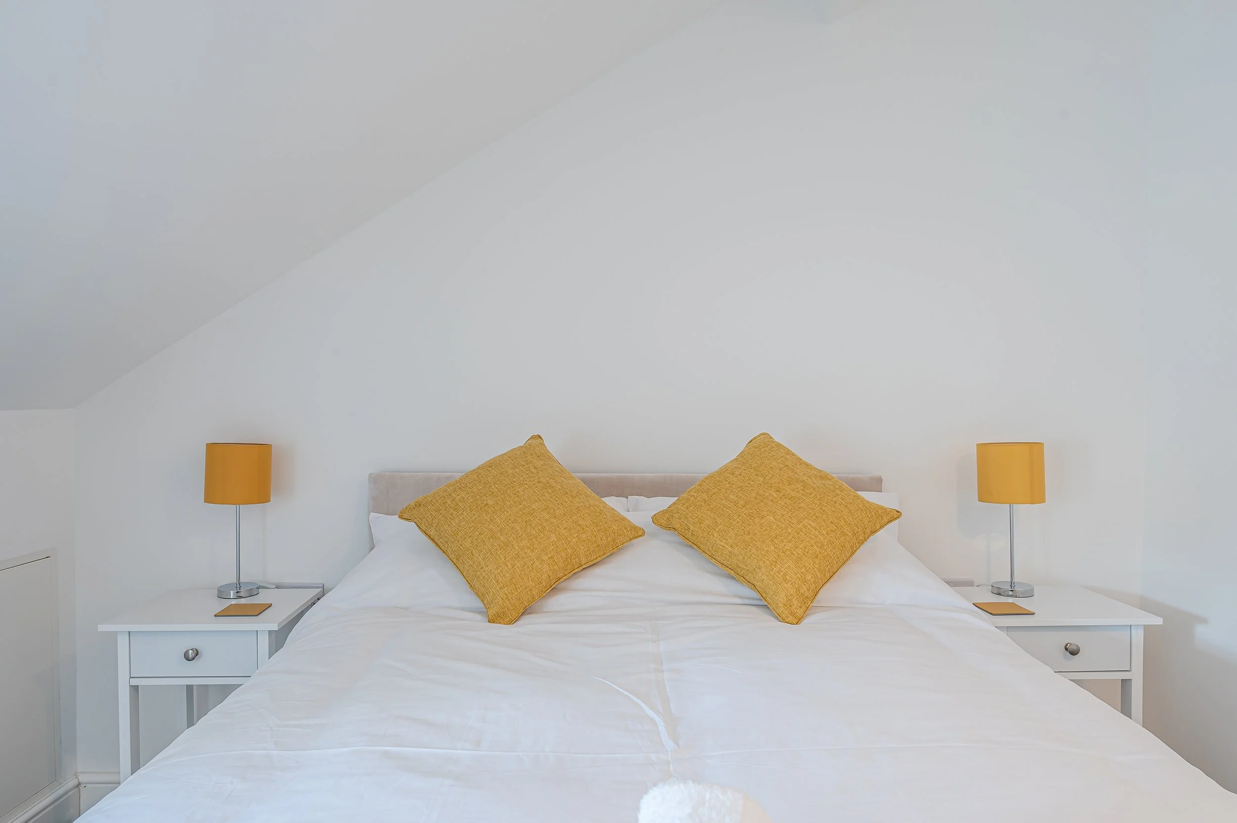 A neatly made bed with white bedding and three mustard-colored pillows, flanked by two white nightstands with matching yellow lamps, set against a white wall with a sloped ceiling.