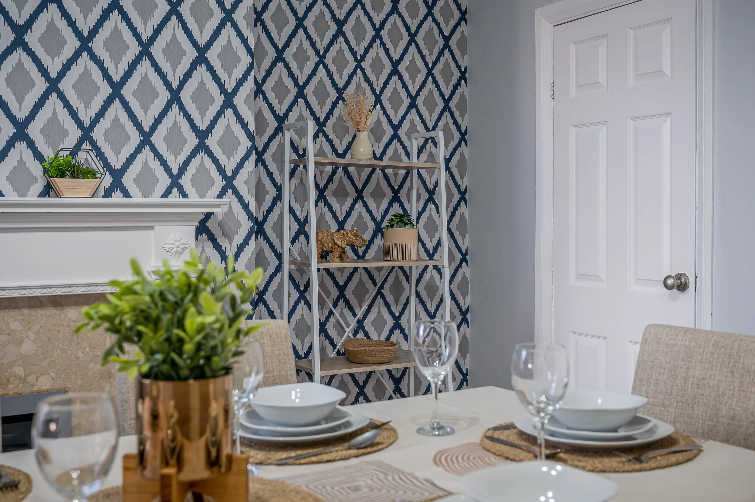 A dining room with a patterned navy and gray wall, a white shelf holding a potted plant and a decorative elephant, a white door, and a table set with white bowls, drinking glasses, and woven placemats with a centerpiece of green plants in copper cont