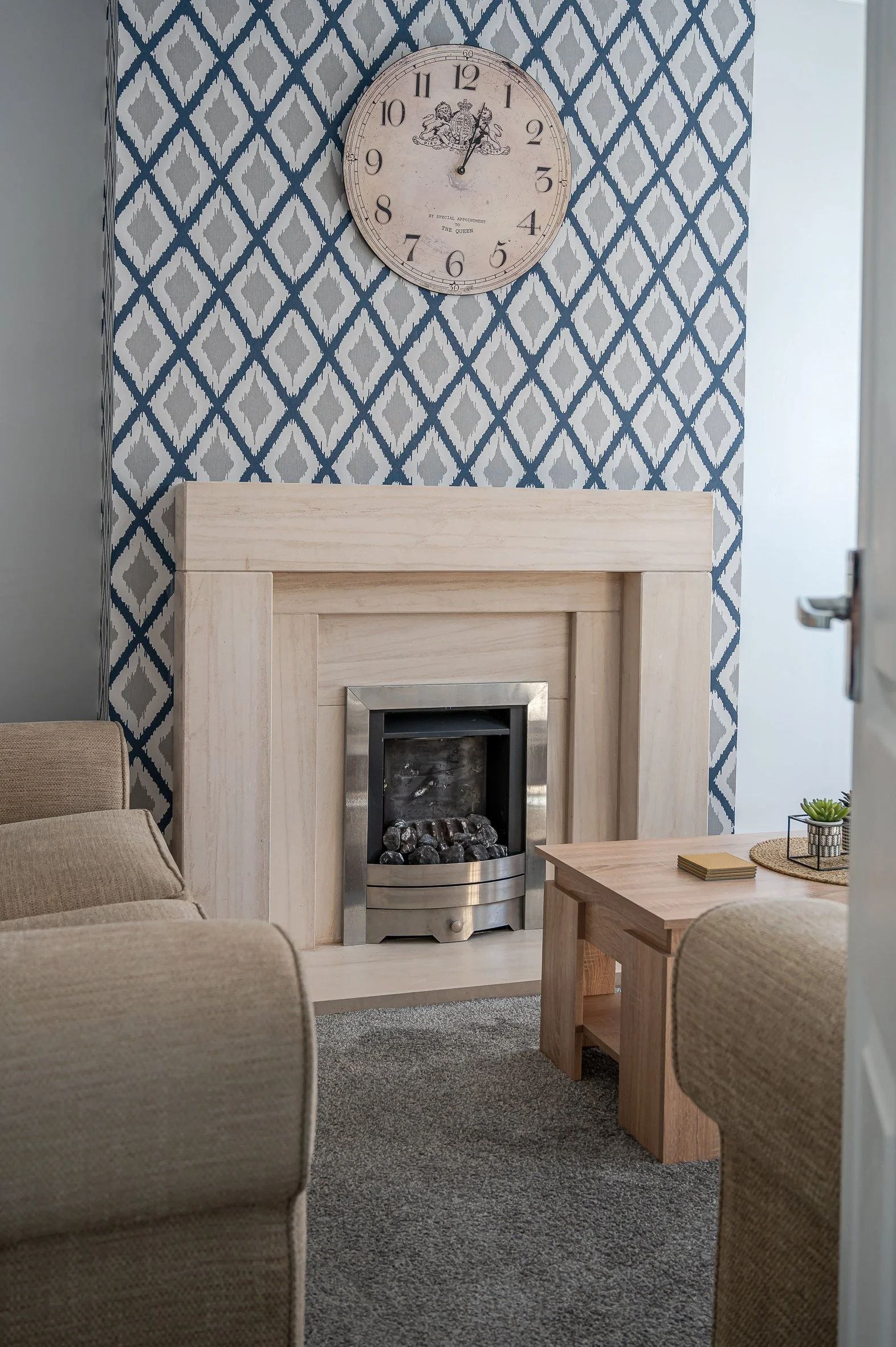Living room with a beige sofa, a wooden coffee table with decoration and books, a fireplace with a metal frame, a wall clock with a crest and text, and patterned blue and white wallpaper.