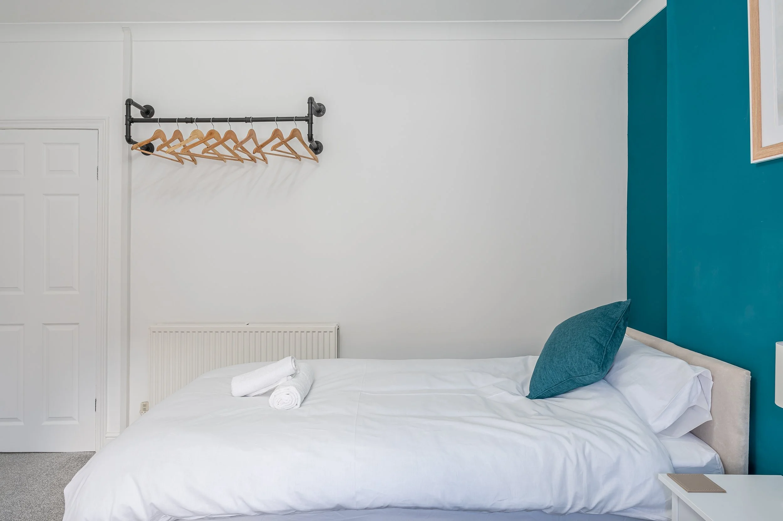 A neatly made bed with white bedding, a teal pillow, and towel rolls, in a modern bedroom with white and teal walls, a framed picture, and a white side table.