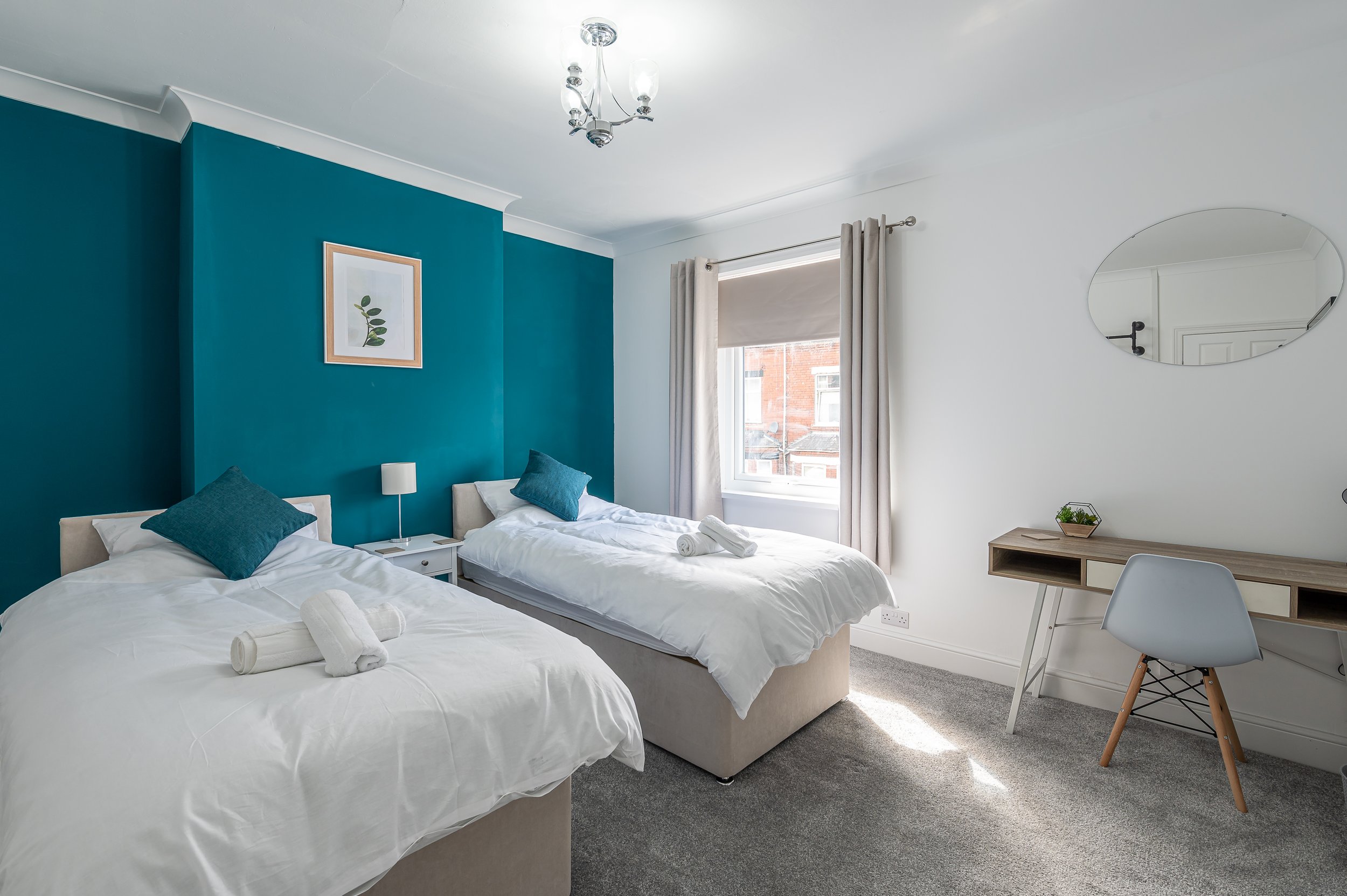 A modern bedroom with two single beds with white bedding and teal pillows. The wall behind the beds is painted teal, with a framed botanical print hanging on it. There is a small white bedside table with a lamp between the beds. A window with cream c