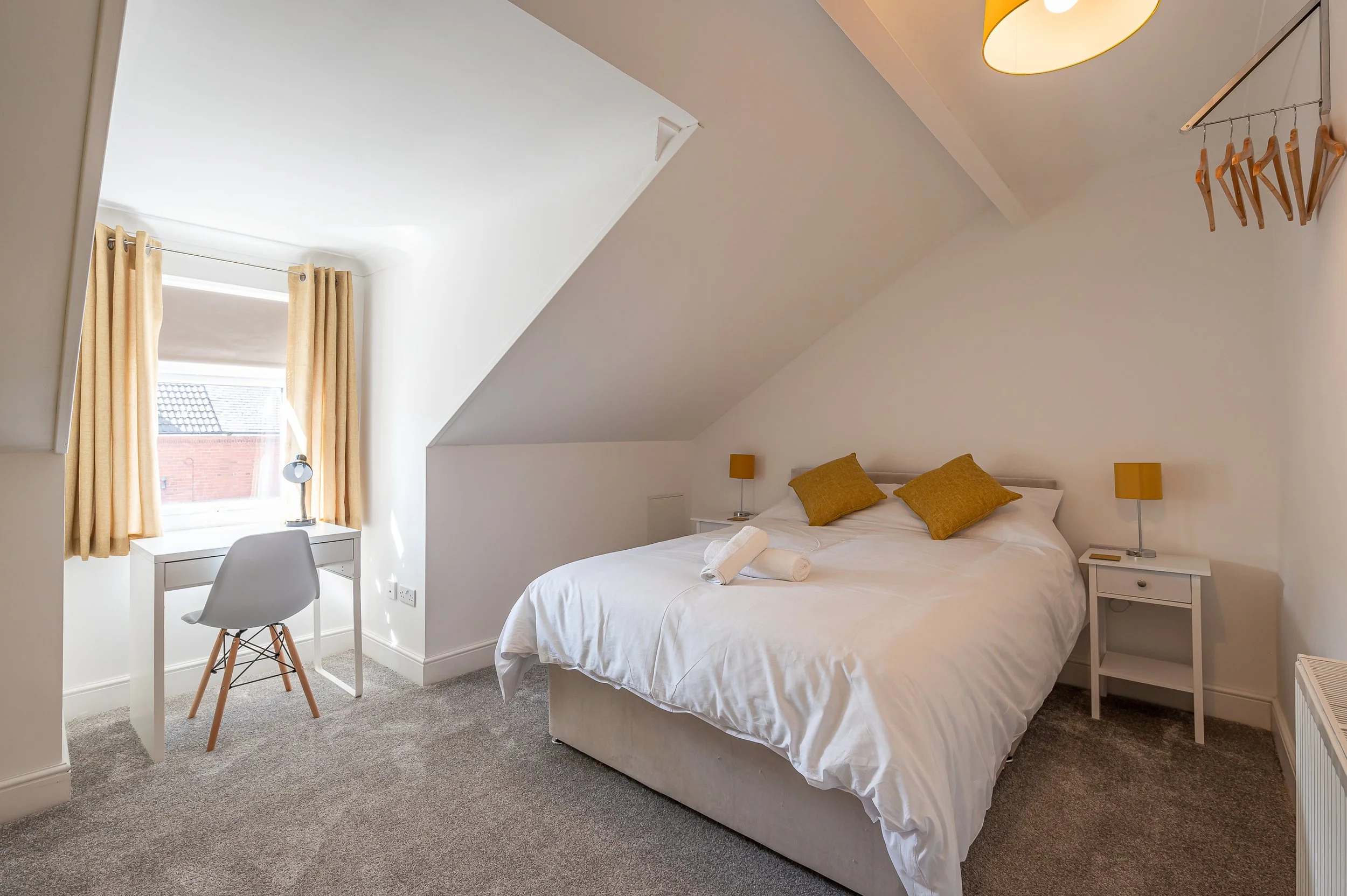 A bright bedroom with a sloped ceiling, featuring a white bed with yellow pillows, bedside tables with yellow lamps, a small white desk with a white chair near a window with yellow curtains, and a hanging clothes rack with wooden hangers.