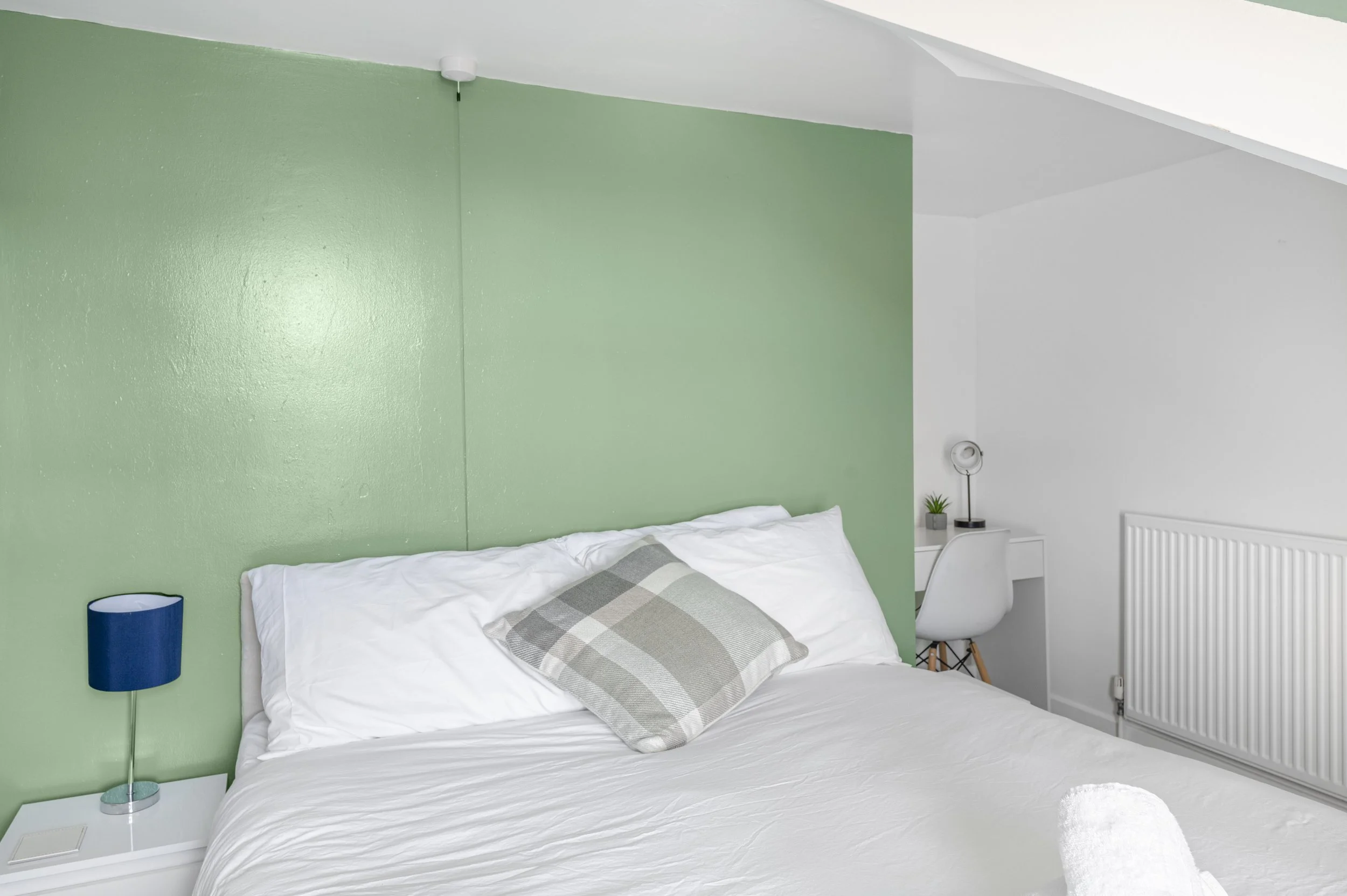 A bedroom with a white bed, green accent wall, white side table with a blue lamp, and a white chair beside a desk with a small plant. There is a white radiator and a spherical wall light.