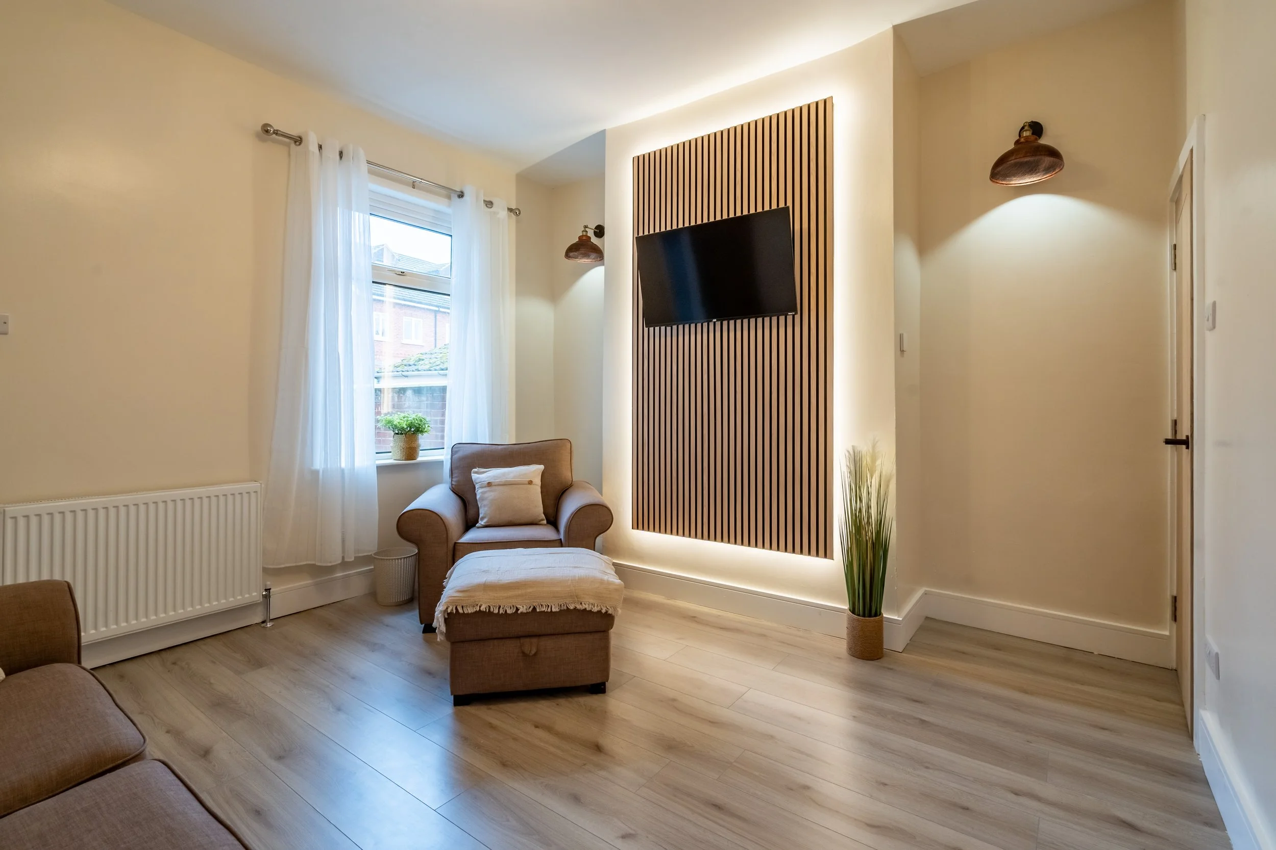 Living room with a beige armchair and matching ottoman, a window with white curtains, a potted plant on the windowsill, a wall-mounted flat-screen TV on a wooden panel, and a tall potted plant in a basket, with hardwood floors and cream-colored walls.