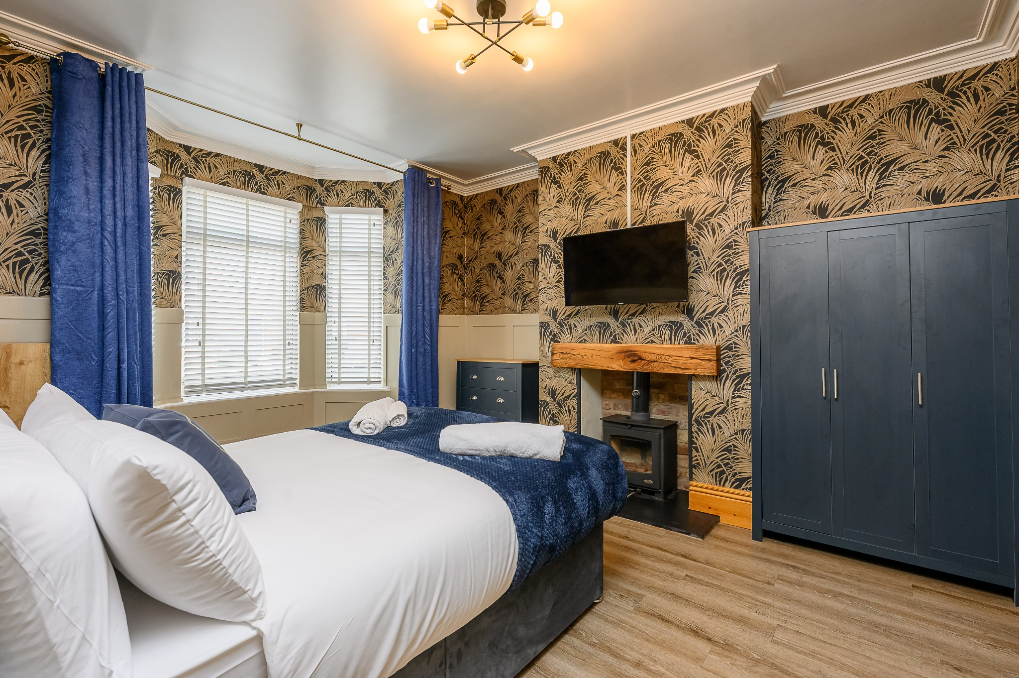A bedroom with white bedding, blue accents, and tropical-patterned wallpaper. There are three windows with white blinds and blue curtains, a mounted flat-screen TV, and a blue wardrobe. The room has a wooden floor and modern ceiling light fixtures.