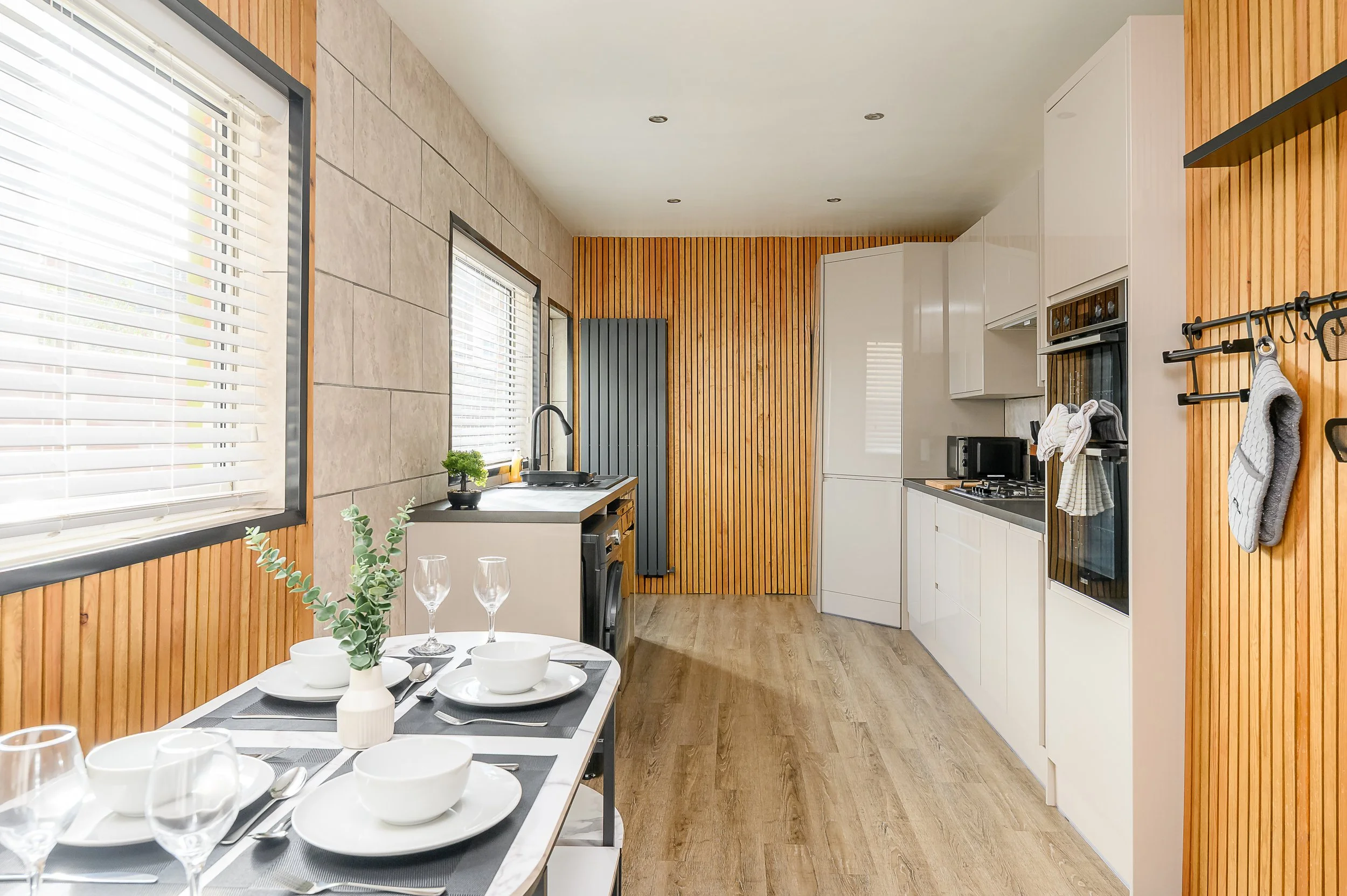 A modern kitchen with white cabinets, wooden wall panels, and a small dining table set for a meal with white dishes and glasses. There are large windows with blinds, a sink, and a stove visible.