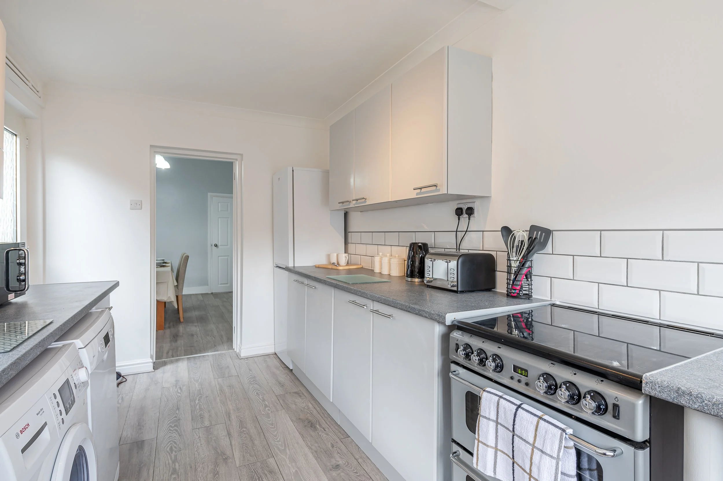 A modern kitchen with white cabinets and a gray countertop, appliances including a stove, toaster, microwave, and washing machines, and a view into a dining area with a table and chairs.