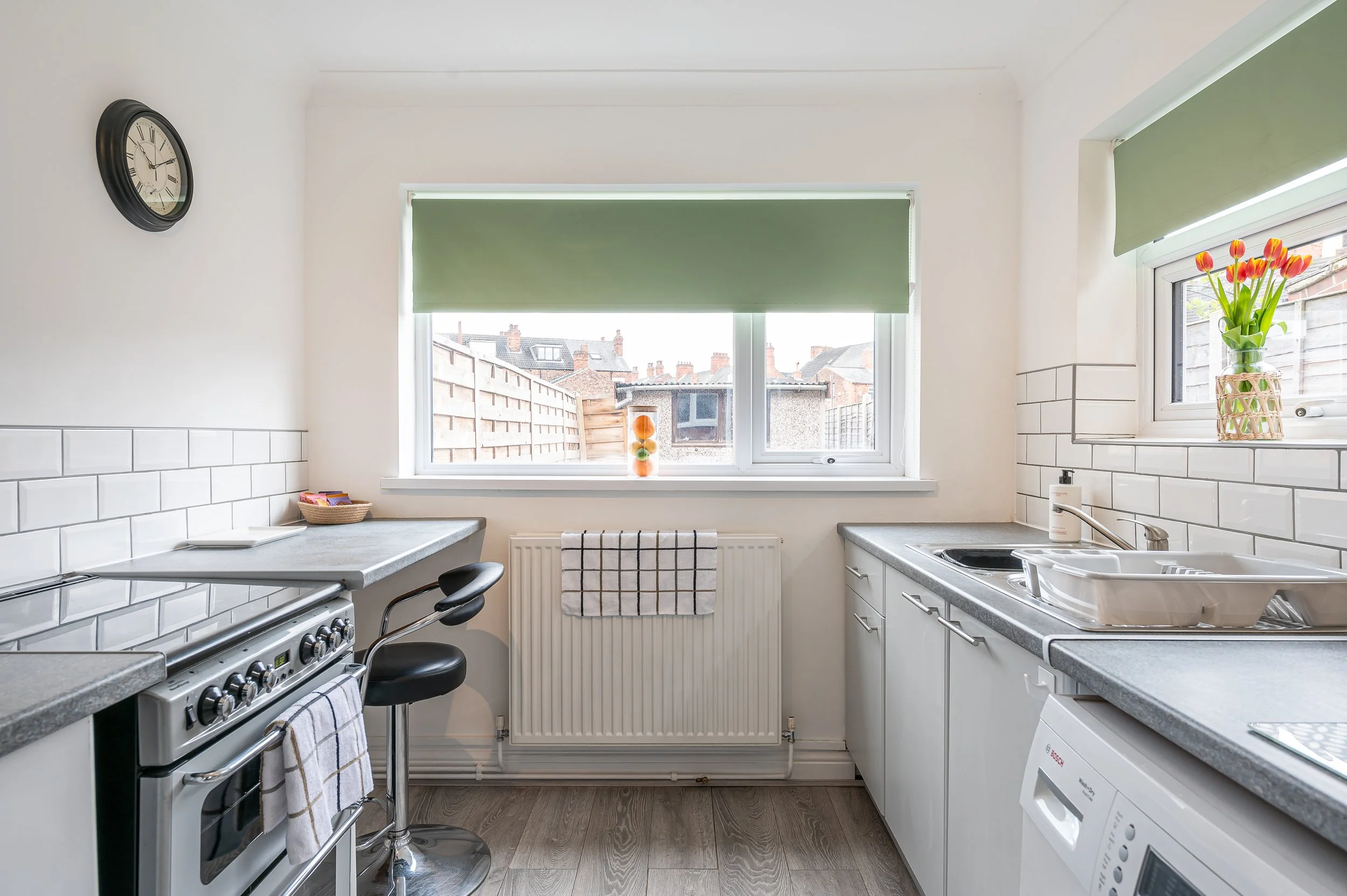 View of a bright kitchen with white cabinets, gray countertops, a stove, a washbasin, a window with green blinds, a vase with tulips, a clock on the wall, and a stool.