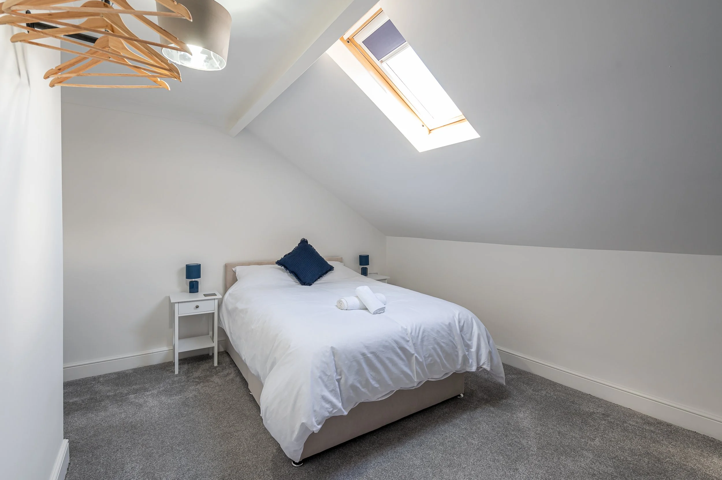 A bedroom with a sloped ceiling, featuring a bed with white linens and two rolled towels on top. There are two navy blue lamps on white nightstands beside the bed. A large skylight window is set into the ceiling, allowing natural light to illuminate 