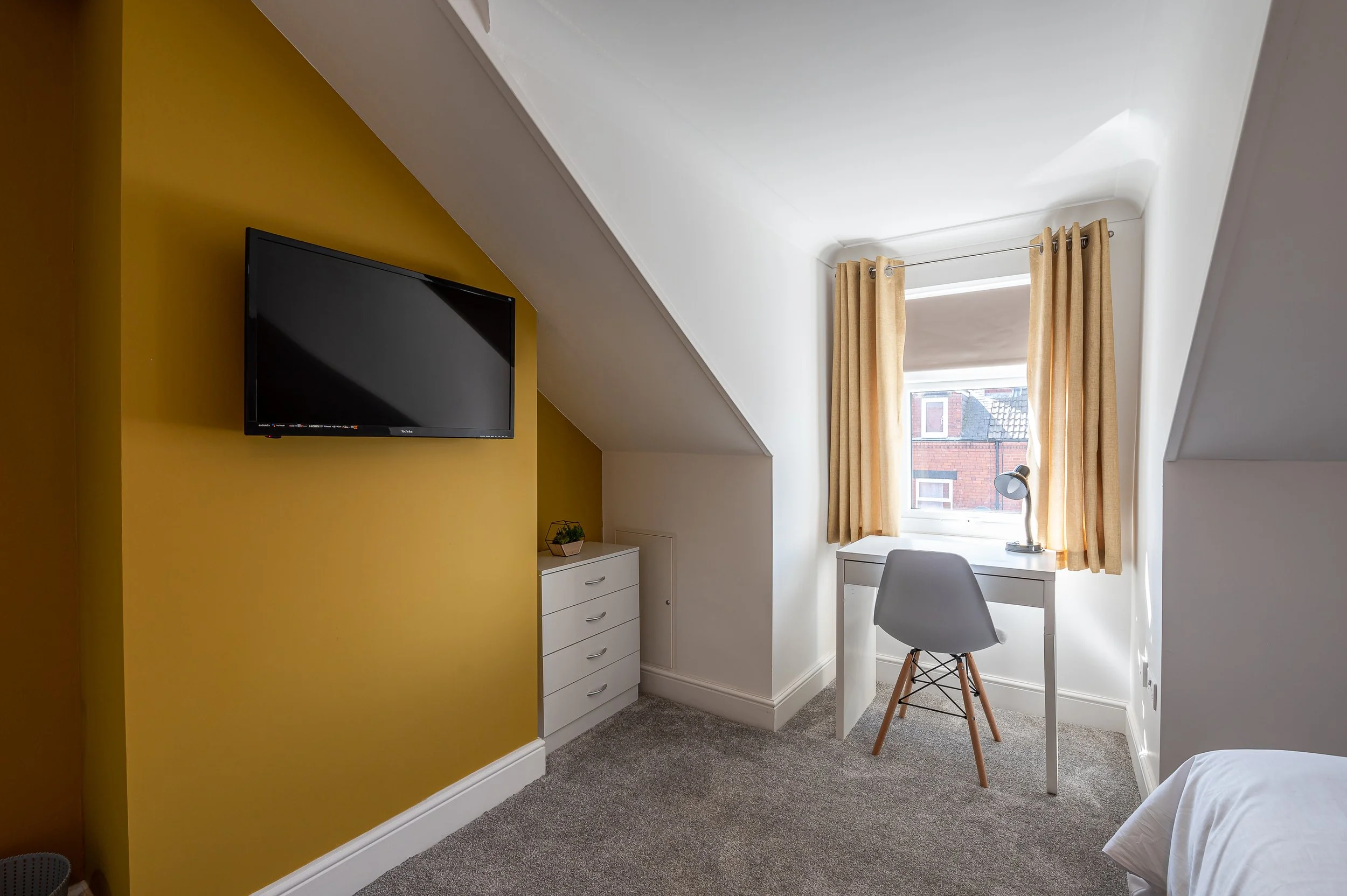 A small room with a white desk, gray chair, a television mounted on a yellow wall, a white dresser, a window with beige curtains, and a lamp.